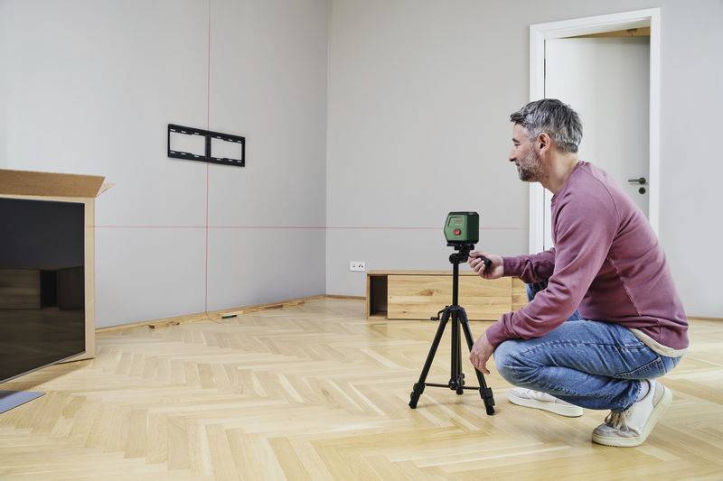 Un homme installe un salon à l'aide d'un trépied et d'un appareil laser. Des lignes de mesure pour la pose sont visibles sur le mur.