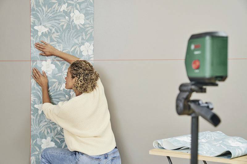Une personne fixe du papier peint à motif floral sur un mur, tandis qu'un niveau laser est posé à côté pour garantir des lignes droites. Une table avec d'autres rouleaux de papier peint est visible en arrière-plan.