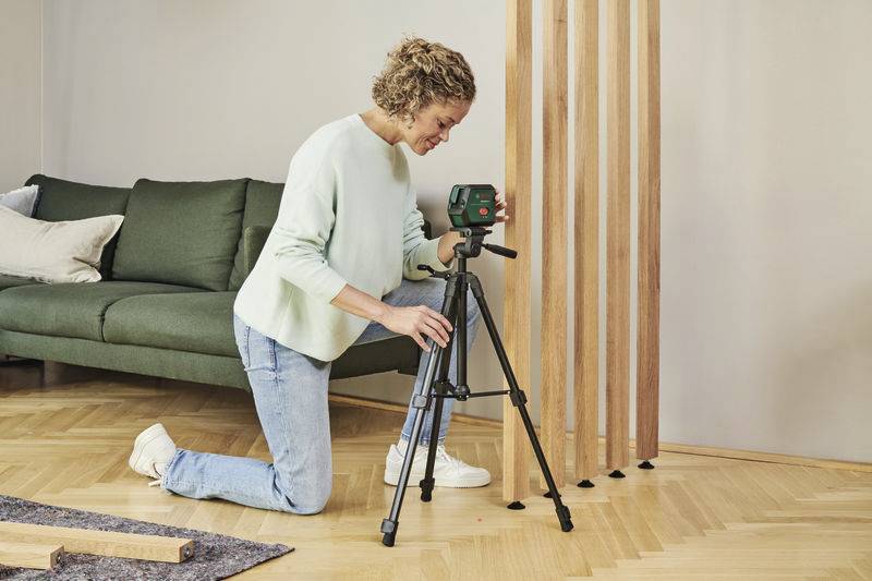 Une personne s'agenouille sur un plancher en bois et ajuste un appareil de mesure sur un trépied dans un salon moderne avec un canapé vert.