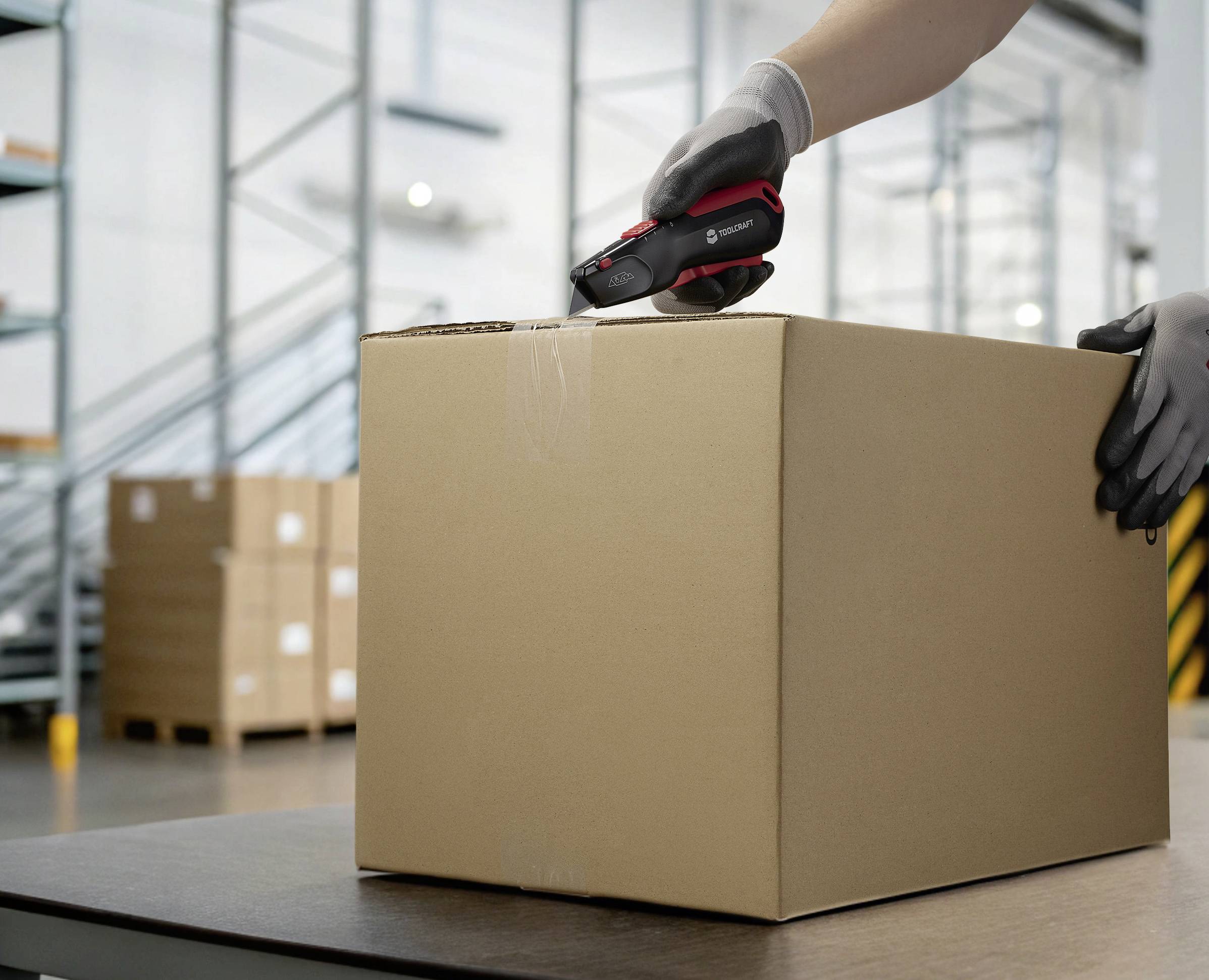 Une personne ouvre un grand carton brun avec un cutter noir et rouge dans un entrepôt avec des étagères.