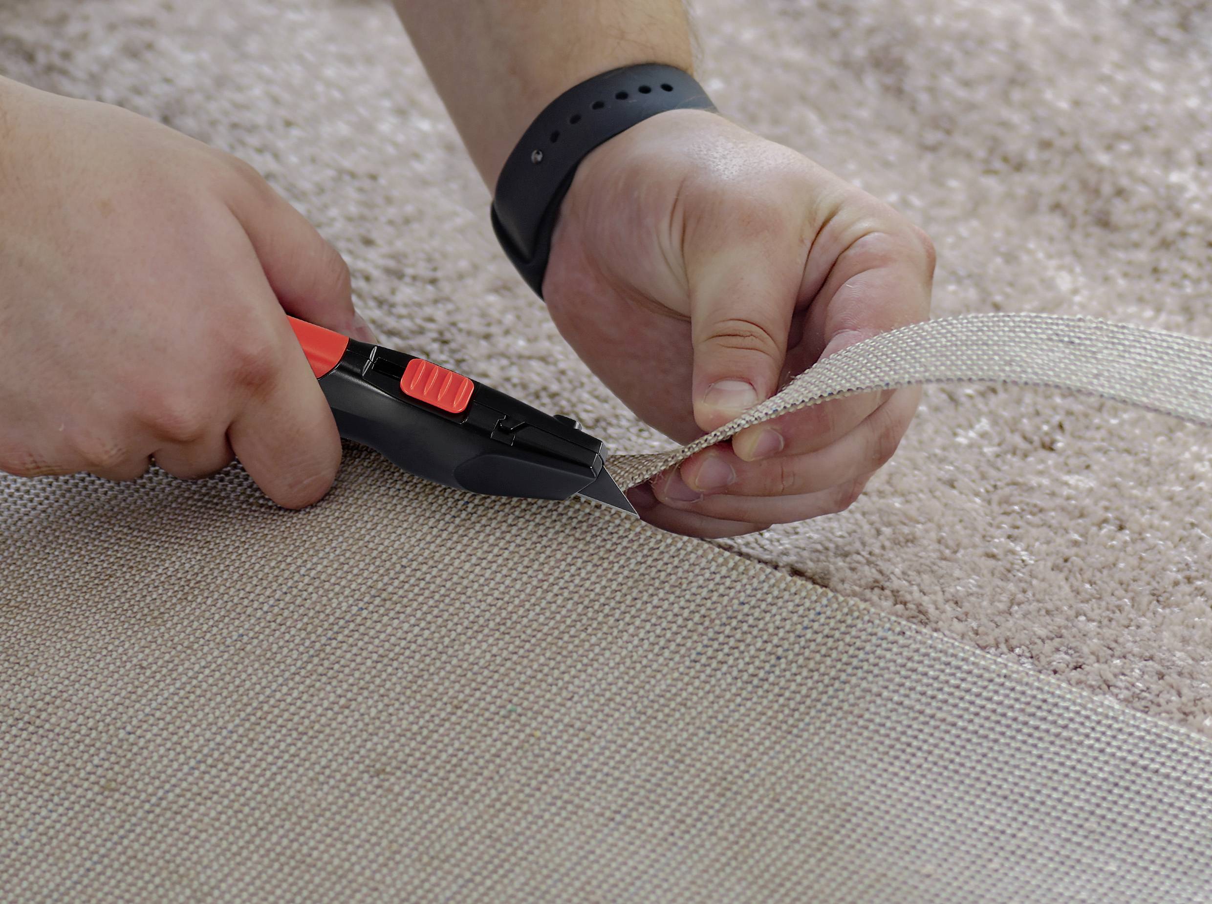 Deux mains découpent un tapis avec un cutter sur un sol beige. Une manchette noire est portée.