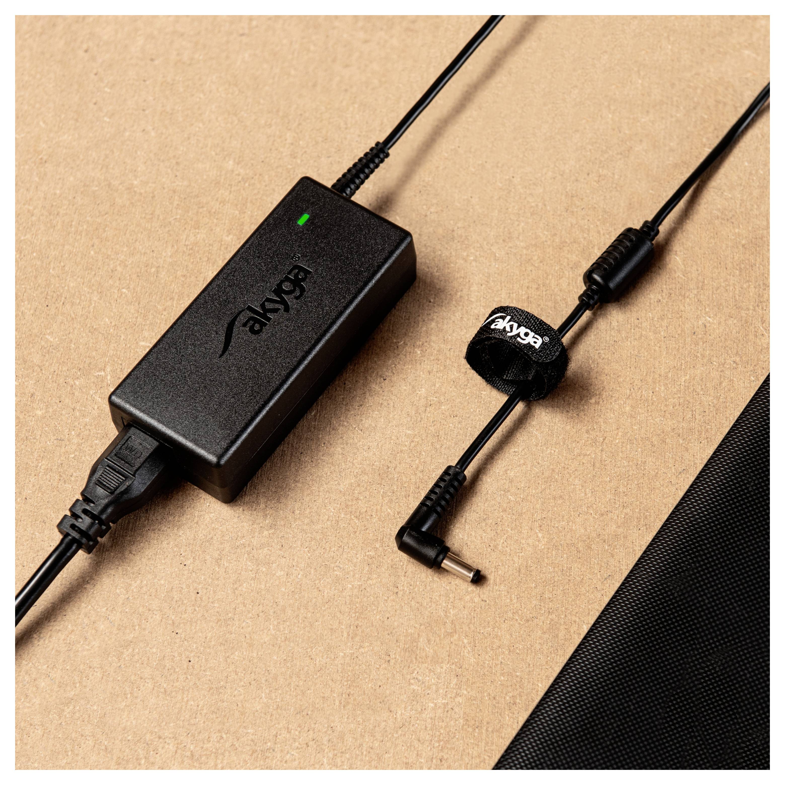 Un chargeur d'ordinateur portable noir avec le logo 'akyga' est posé sur un fond marron à côté d'un câble avec une attache velcro.
