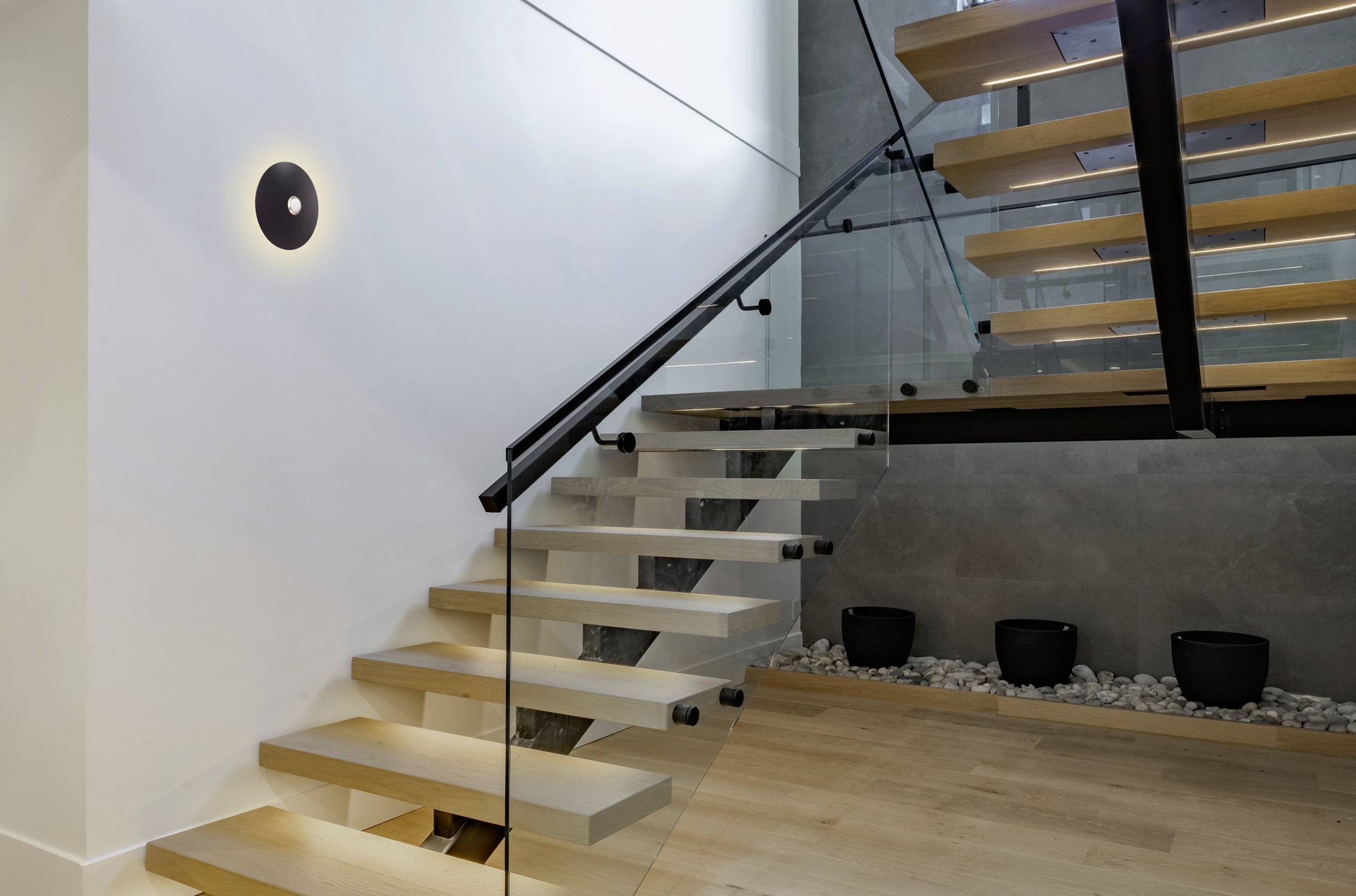 Un escalier moderne ouvert avec des marches en bois et des garde-corps en verre est représenté, avec un luminaire mural élégant et des plantes en pot sur le palier.