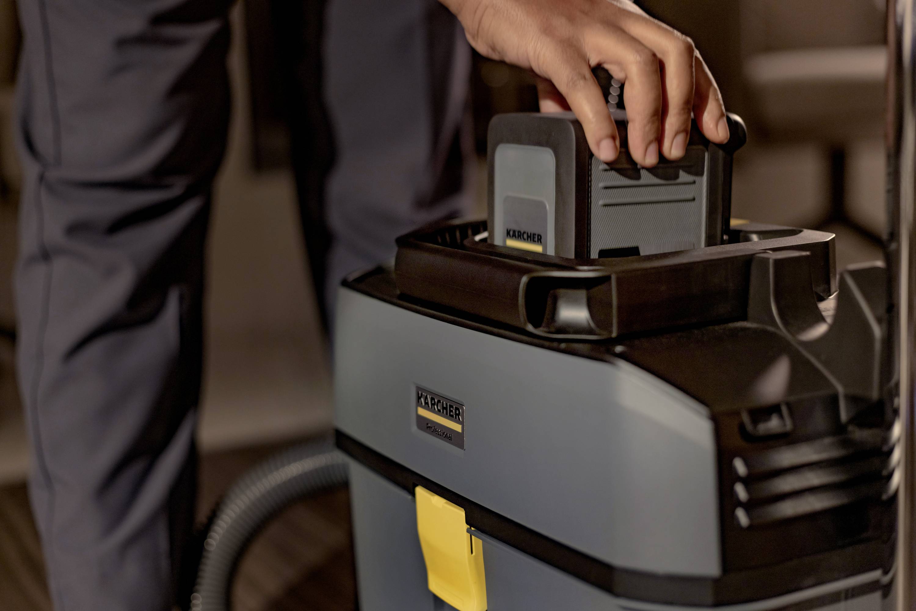 Une personne installe une batterie dans un aspirateur industriel. L'aspirateur est gris avec un élément jaune et présente une construction robuste.