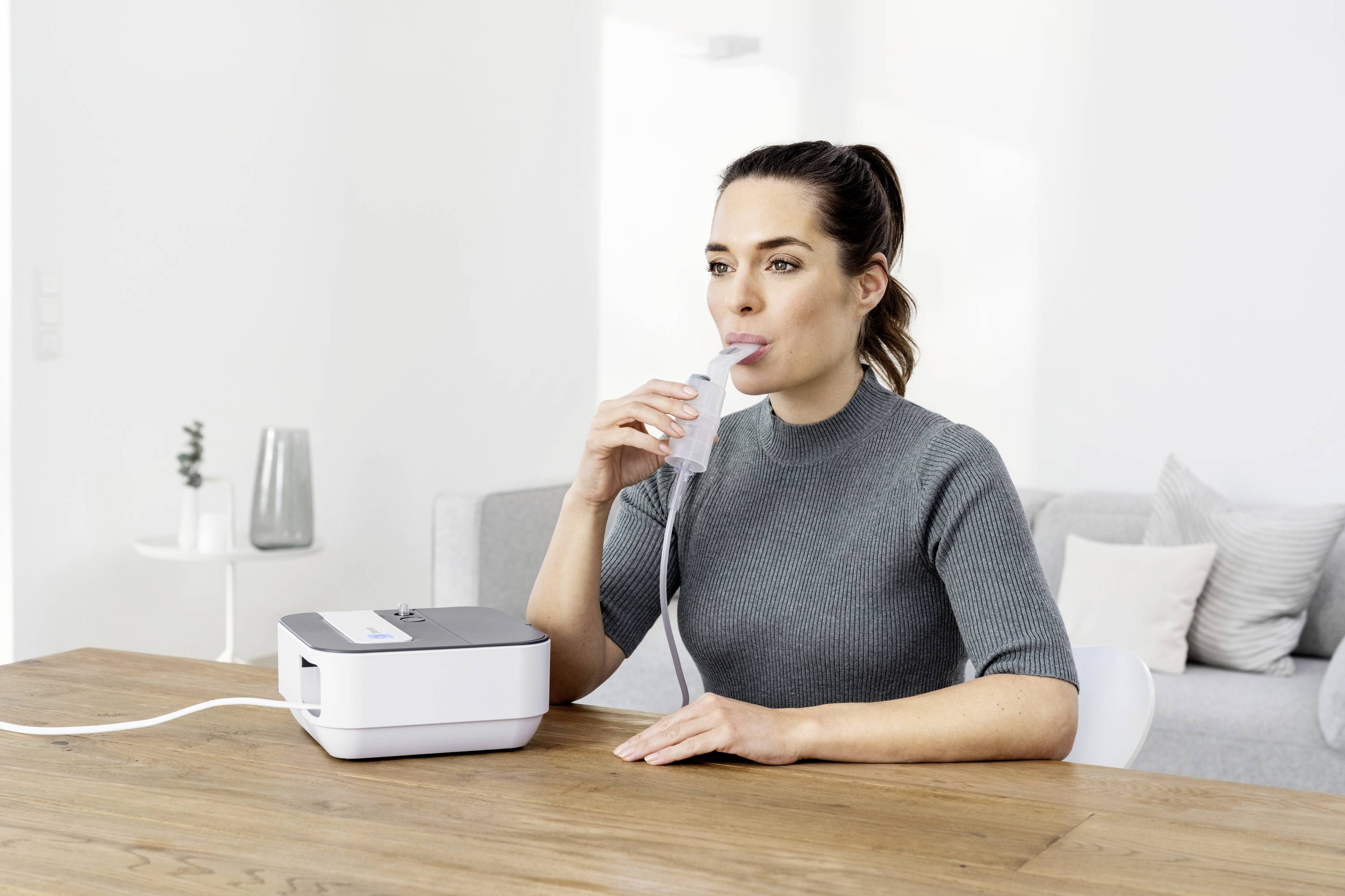 Une femme utilise un appareil d'inhalation, assise à une table en bois dans une pièce moderne et lumineuse.