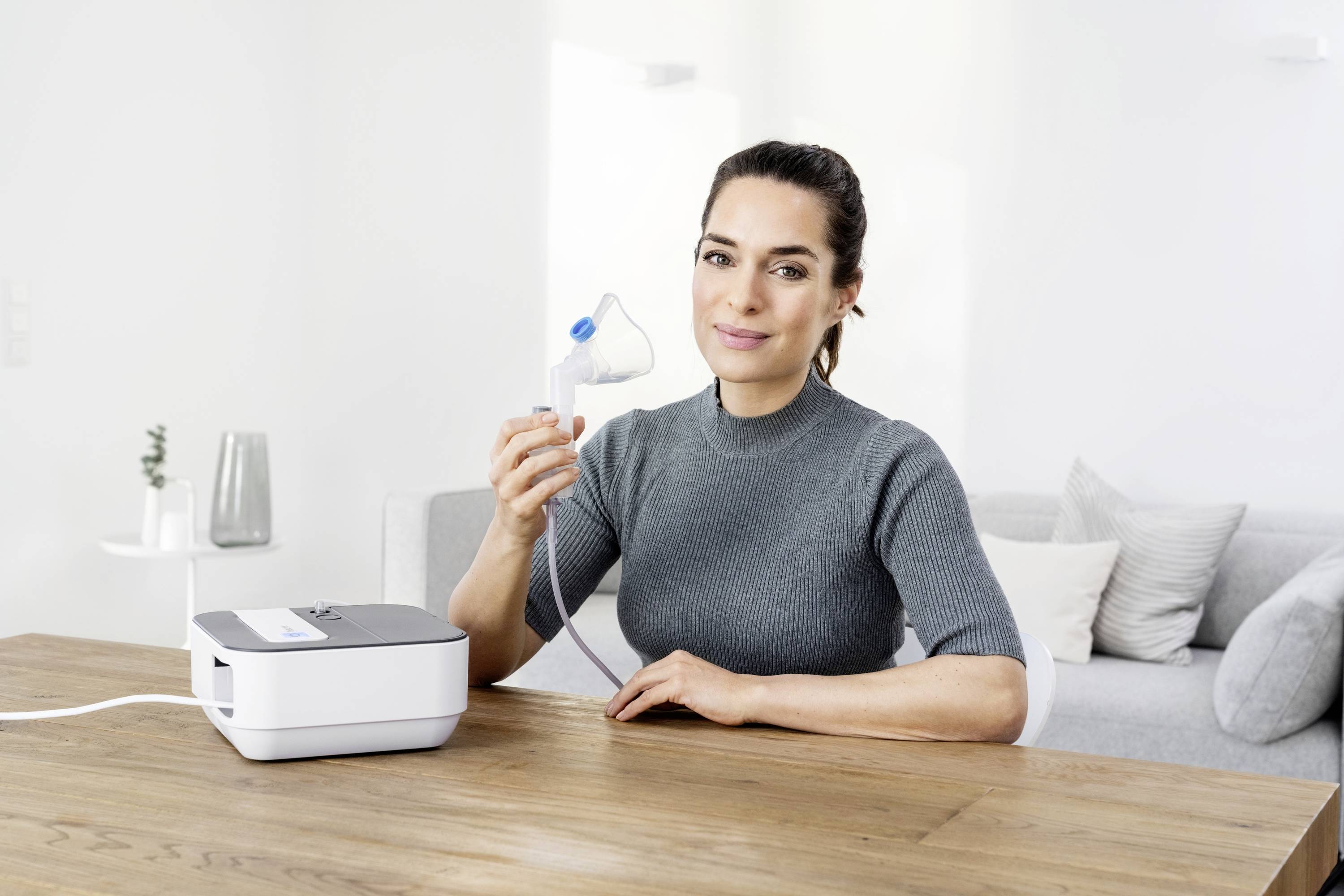 Une personne est assise à table et tient un dispositif d'inhalation. L'appareil est posé à côté d'elle, et en arrière-plan se trouve un environnement de salon lumineux.