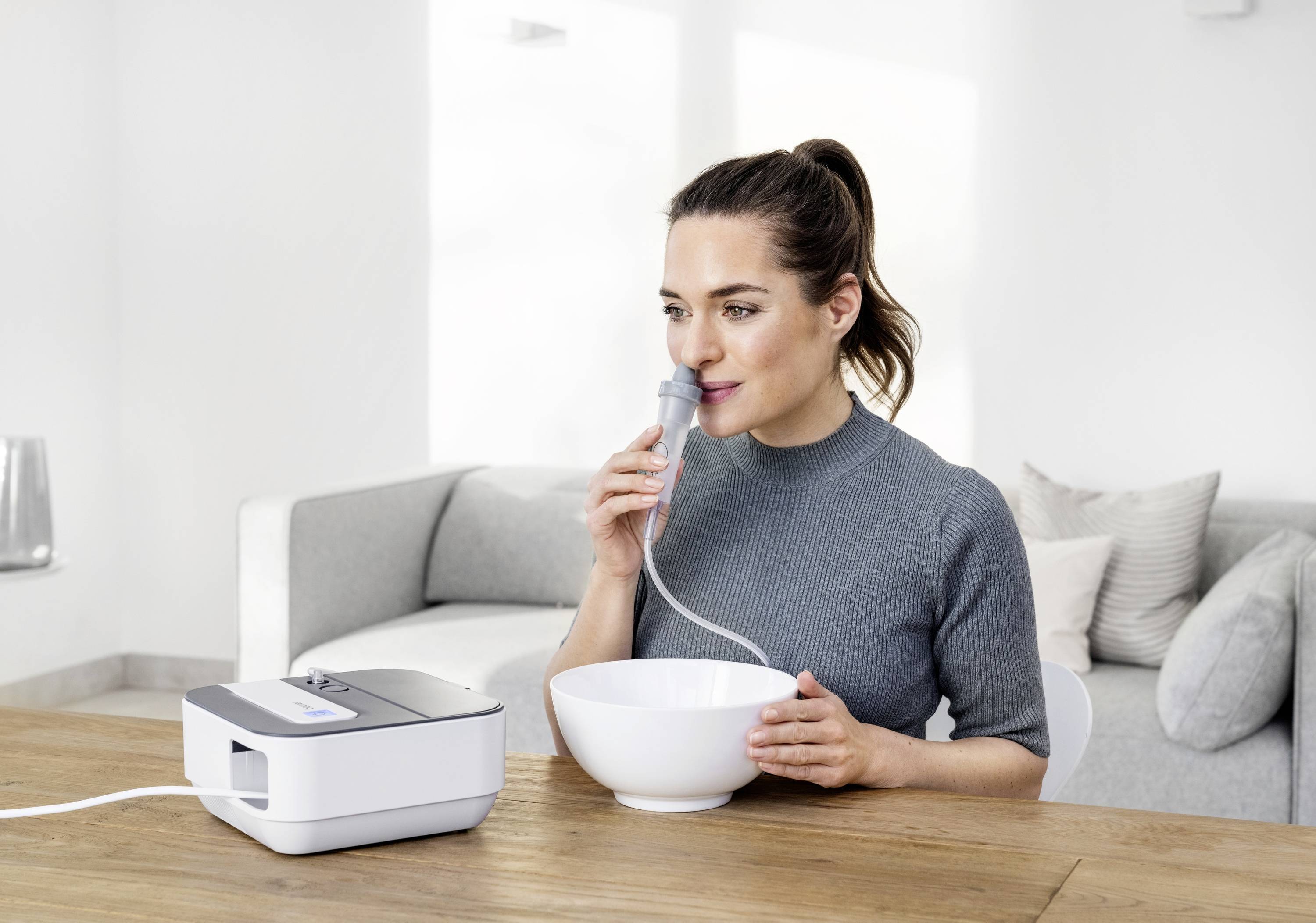 Une femme utilise un appareil d'inhalation à une table. Elle tient un bol et inhale à travers un tube, dans un salon lumineux.