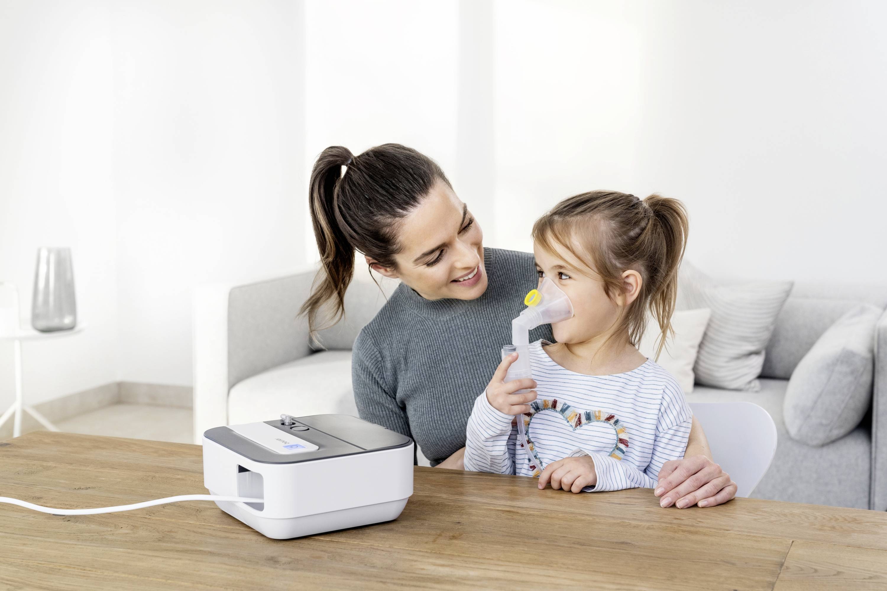 Une femme est assise à une table et regarde avec un sourire un petit fille qui utilise un appareil d'inhalation.