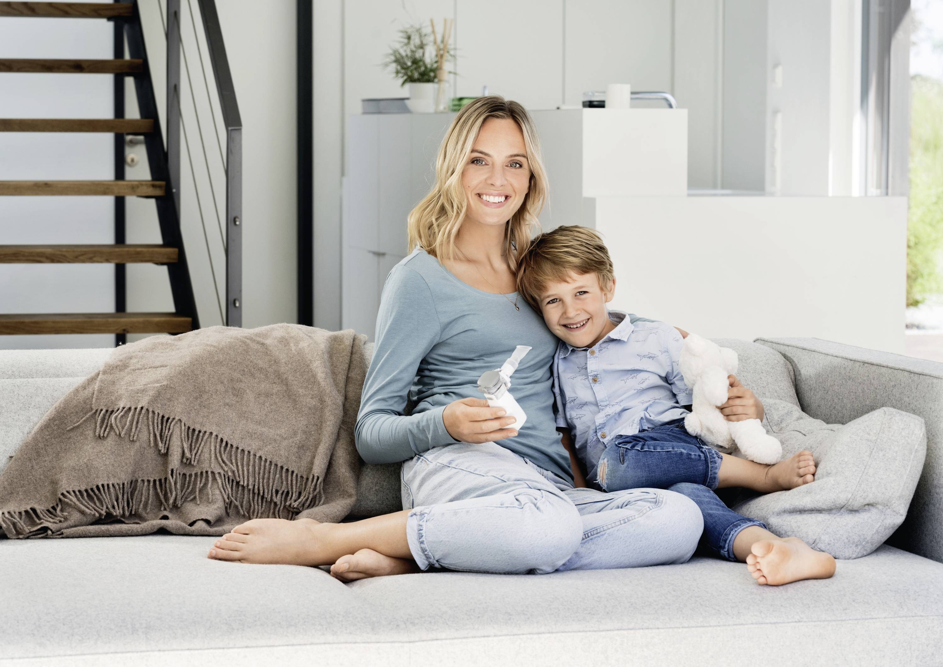 Une femme et un enfant sont assis en souriant sur un canapé gris. La femme tient un doudou, et l'enfant a un animal en peluche dans la main.