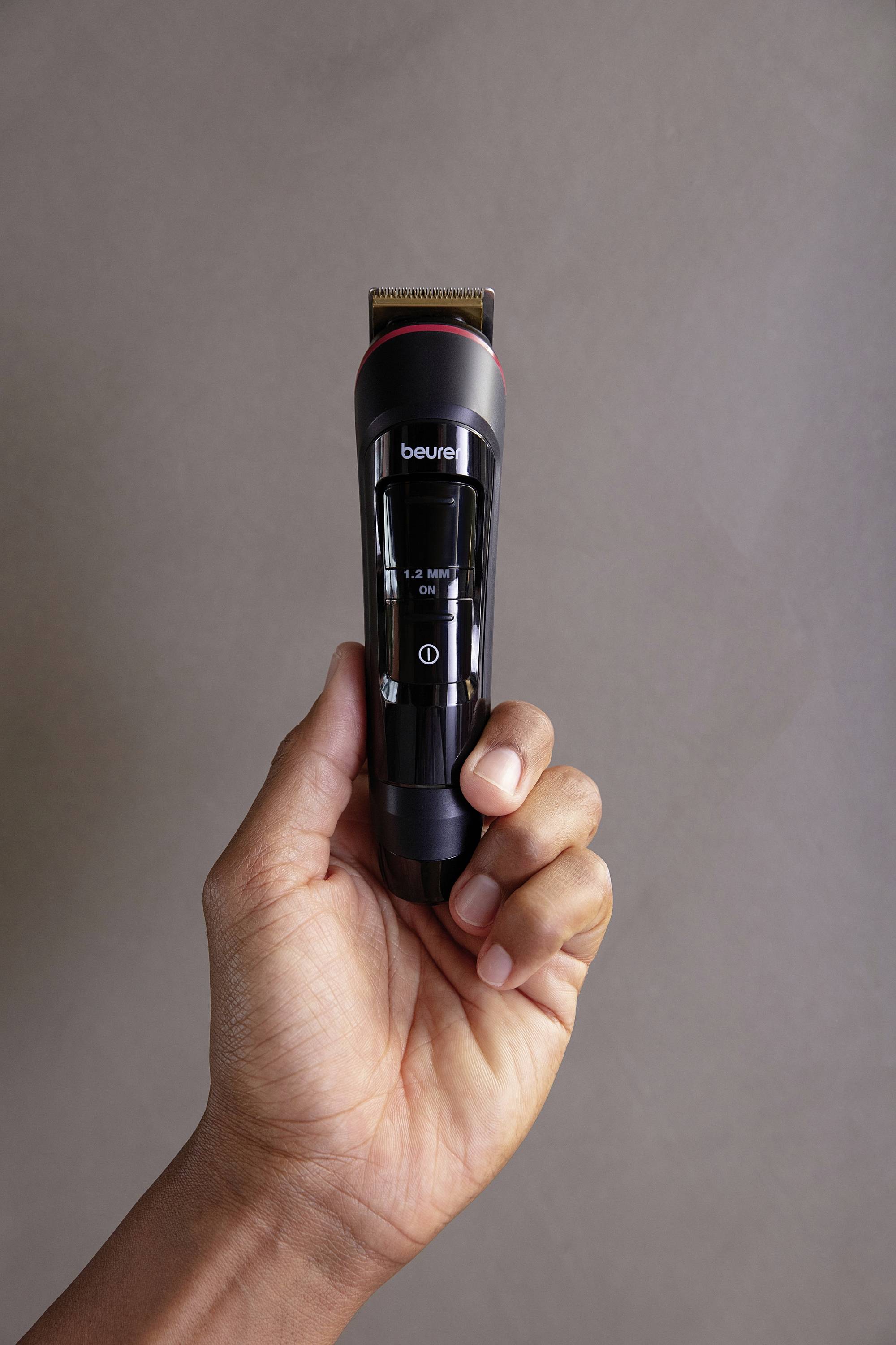 Une main tient un tondeuse électrique noire portant l'inscription 'beurer'. L'arrière-plan est neutre.