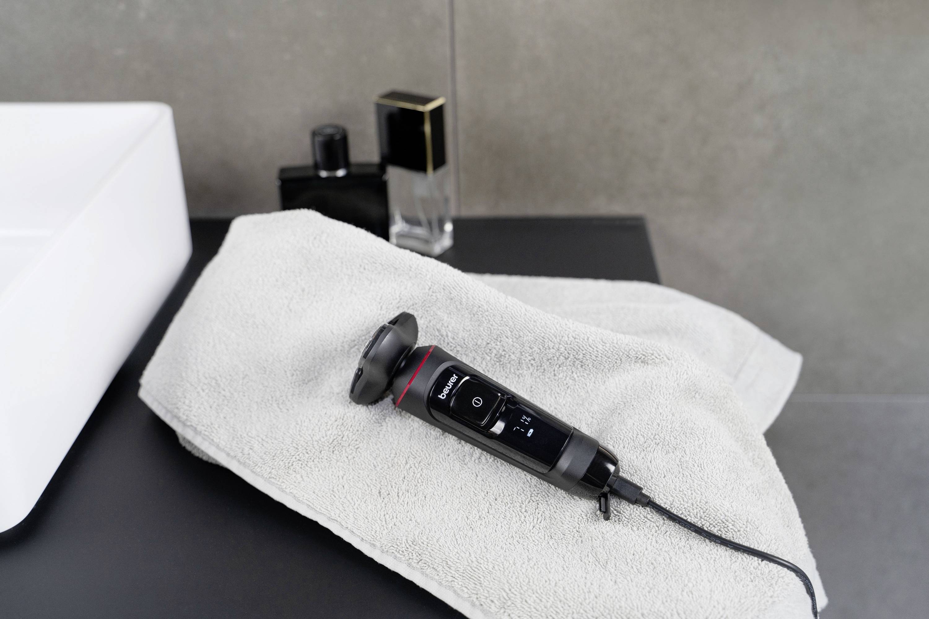 Un tondeuse électrique repose sur une serviette sur un lavabo, entourée de flacons de parfum en arrière-plan.