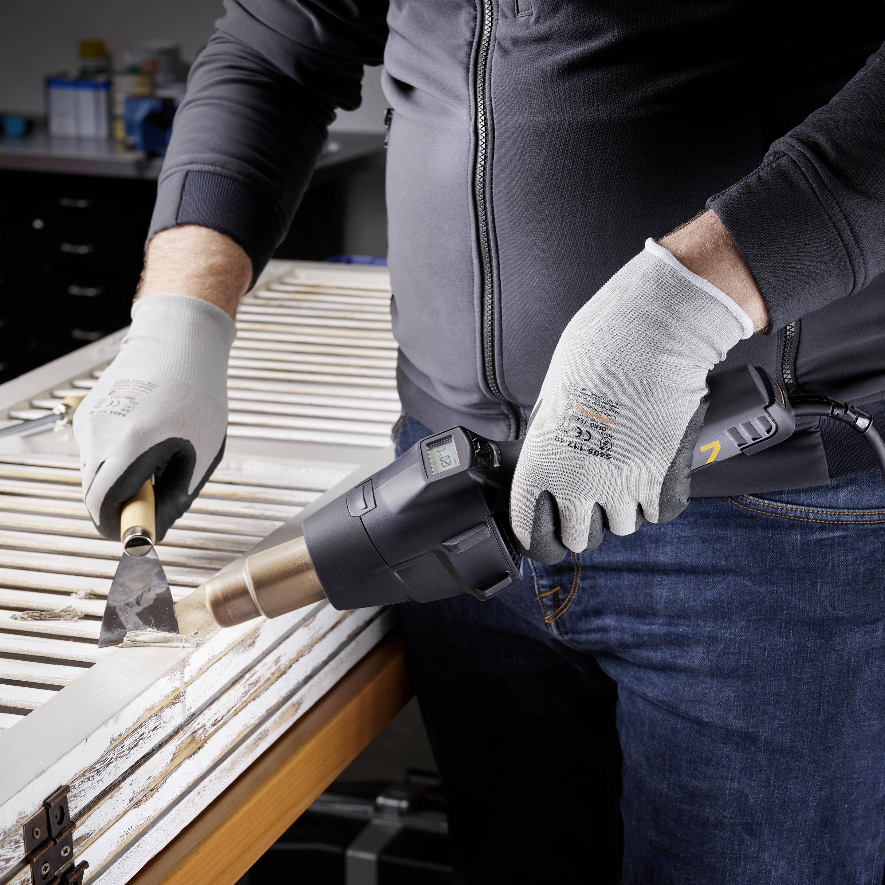 Une personne utilise un décapeur thermique pour enlever une ancienne peinture d'un cadre en bois, tout en tenant une spatule dans l'autre main.