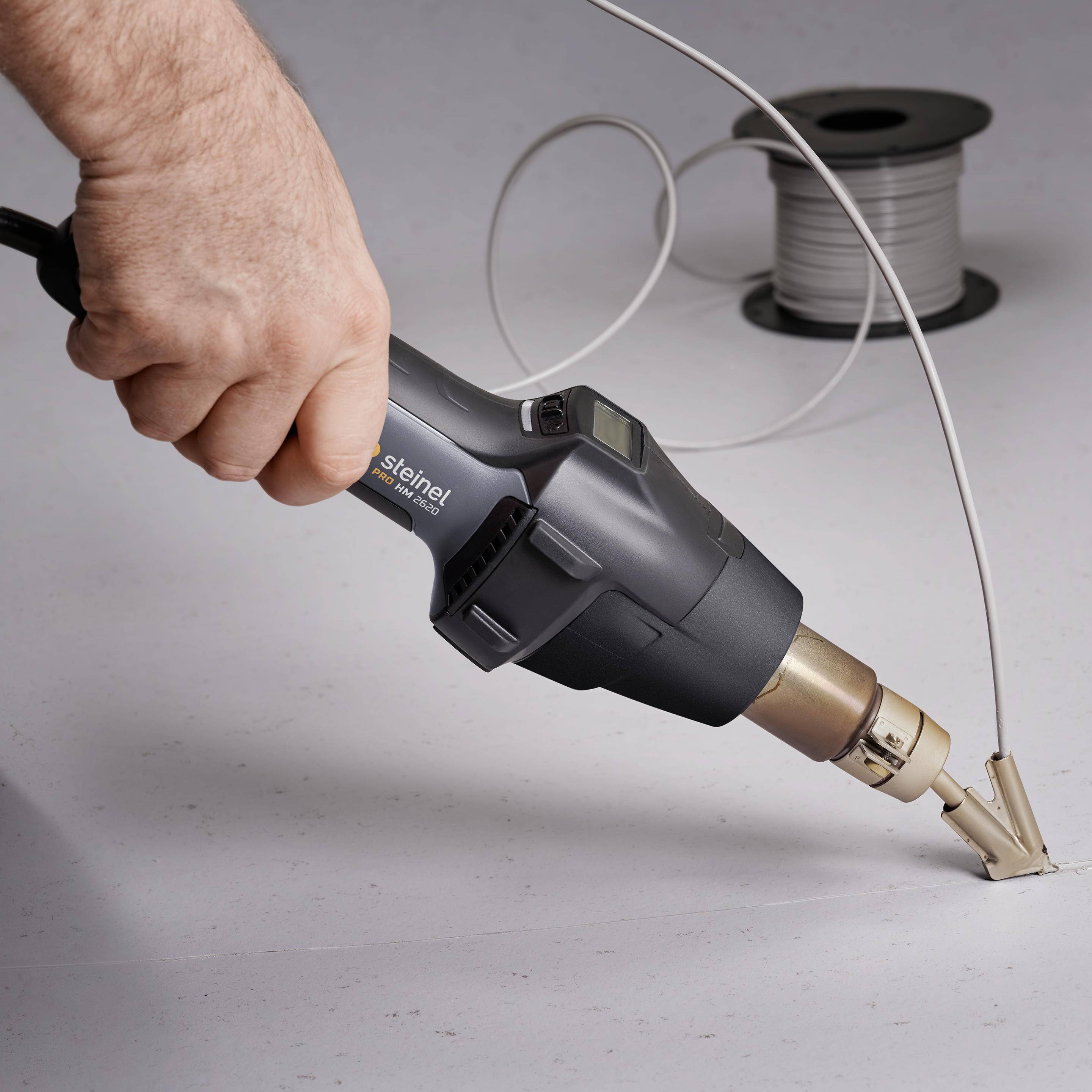 Une main tient un pistolet à air chaud dirigé vers une section de câble pour le rétrécir. En arrière-plan, un rouleau de câble.