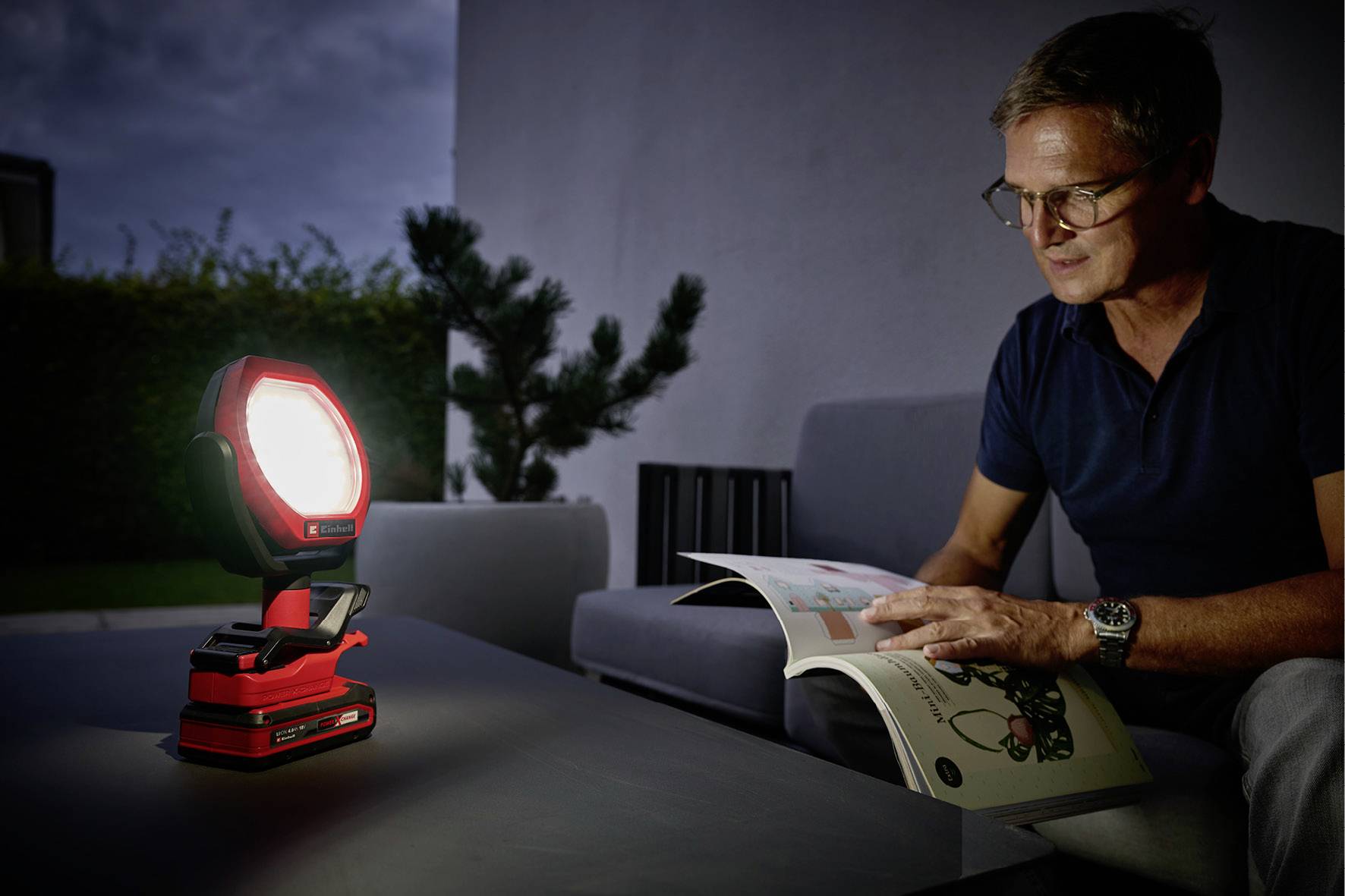 Un homme est assis dans une pièce faiblement éclairée et lit un magazine, tandis qu'une lampe chargée et lumineuse répand sa lumière sur une table.