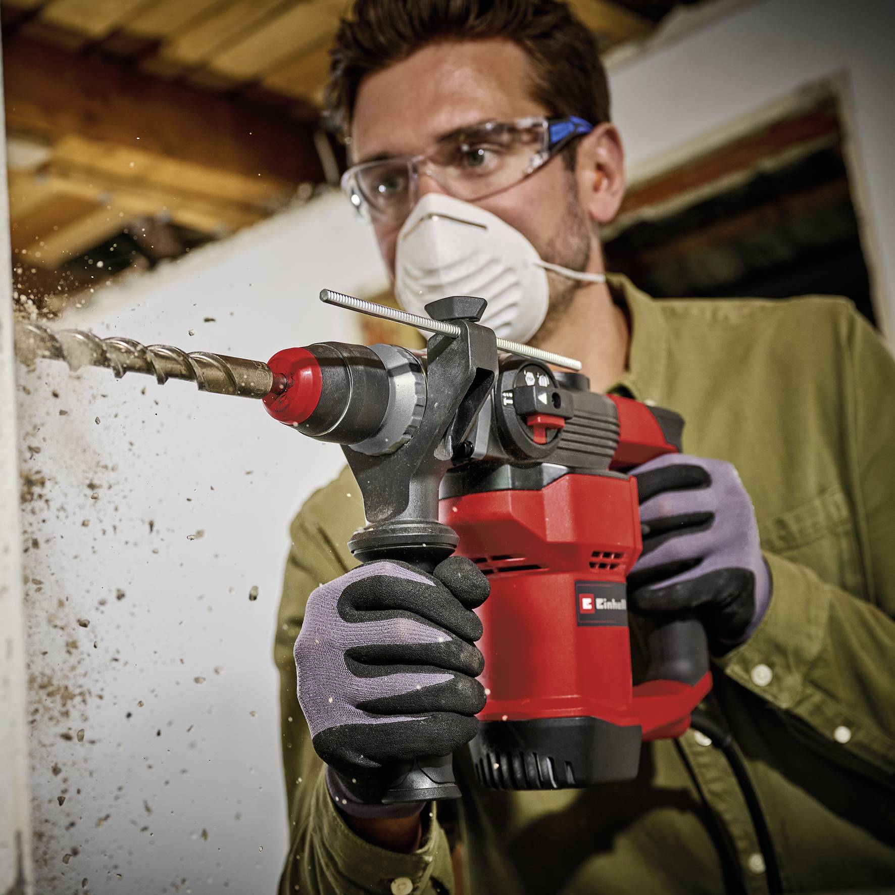 Une personne portant des lunettes de protection fore un mur avec une perceuse à percussion rouge et noire. Elle porte des gants de sécurité.
