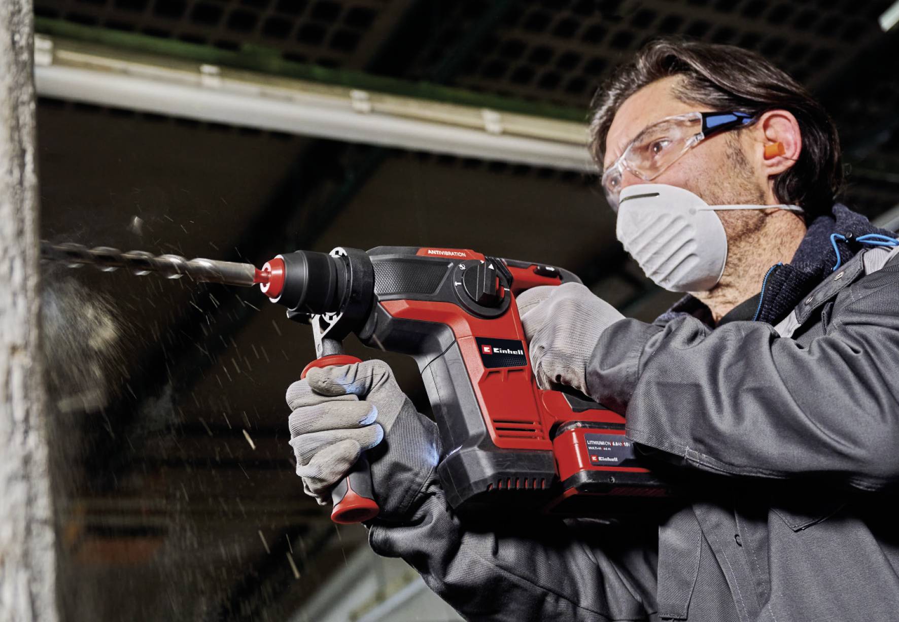Un homme en tenue de protection fore un mur en bois avec une machine-outil électrique rouge. Il travaille de manière concentrée dans un atelier.