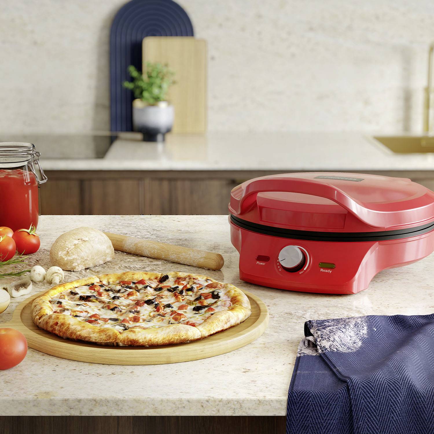 Un fabricant de pizza rouge se trouve sur un plan de travail de cuisine avec une pizza fraîchement cuite. À côté, des ingrédients tels que des tomates et des champignons sont disposés.