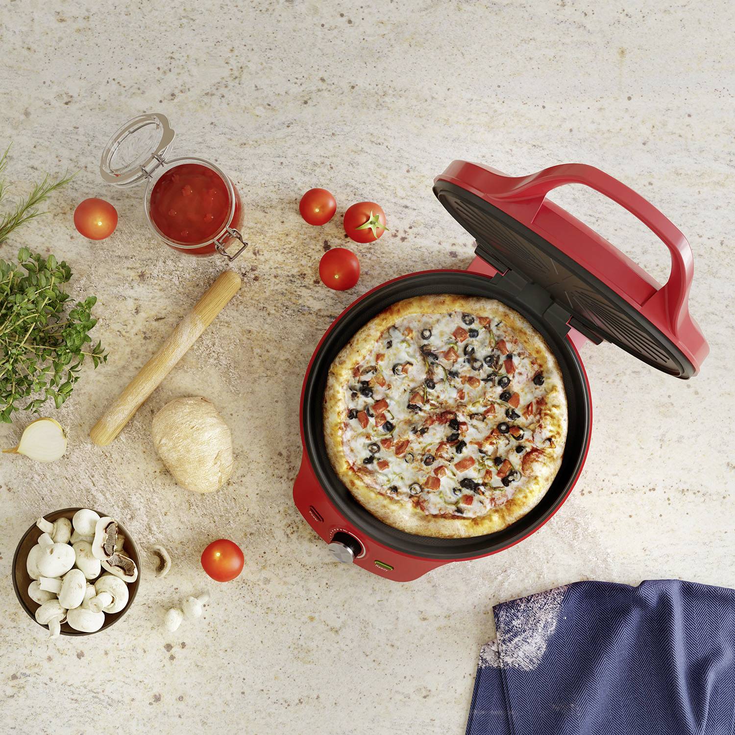 Pizza dans un four rouge à pizza, entourée de tomates, de champignons, d'herbes et d'un verre de sauce tomate sur une table.