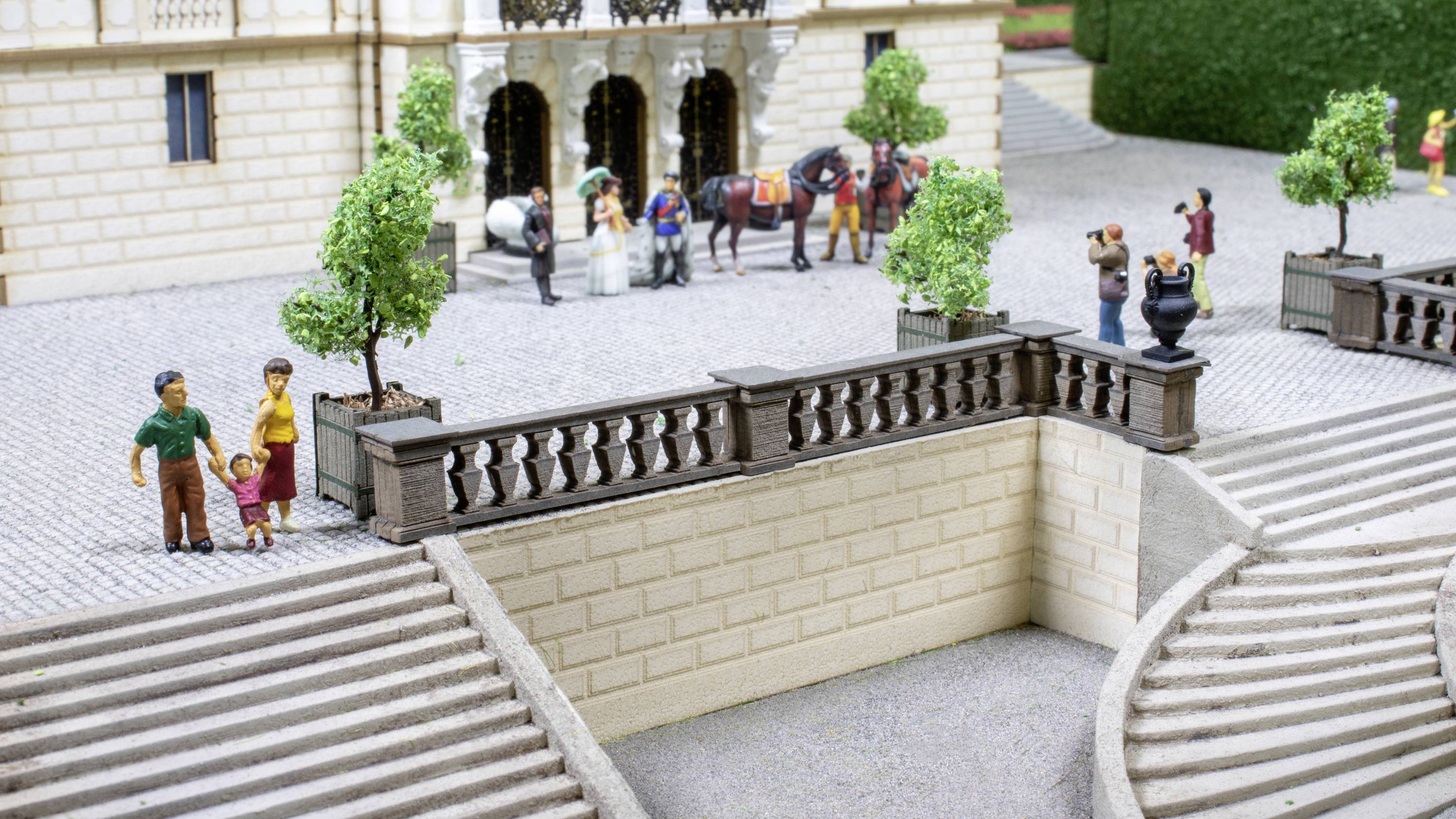 Une scène de modèle miniature montre des personnes dans une cour devant un bâtiment ; un chariot à cheval et des arbres sont visibles.
