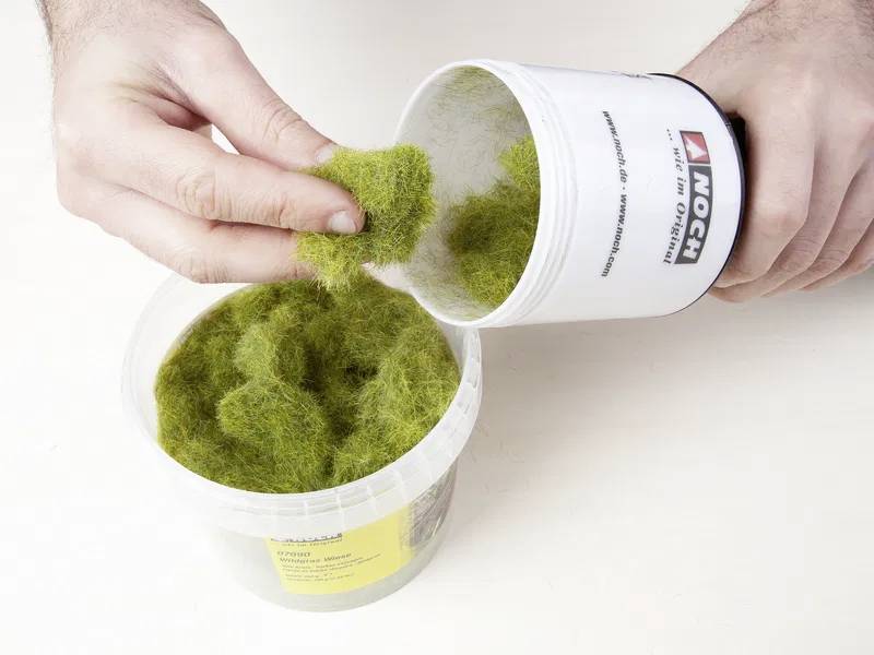 Une personne verse de la fibre synthétique verte imitant l'herbe d'un récipient dans une grande bassine en plastique, utilisée pour des maquettes de paysage ou des dioramas.