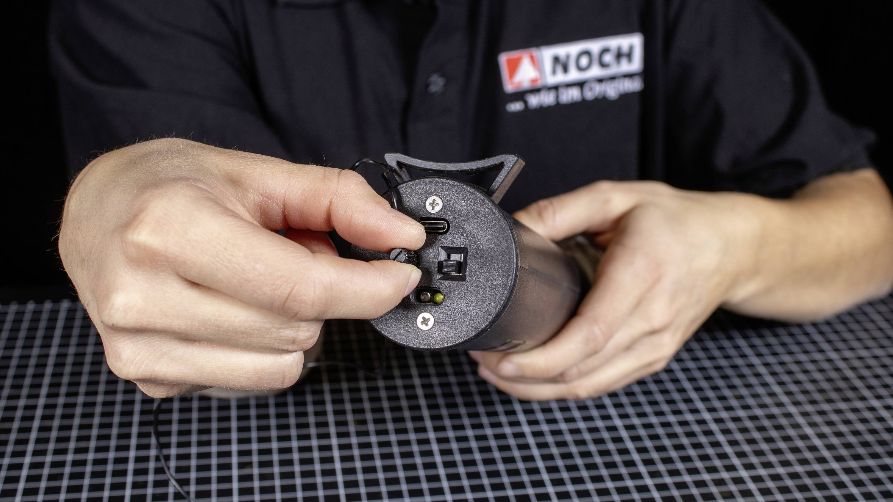 Une personne tient un petit appareil électrique avec des connexions. Un logo est visible sur la chemise de la personne.