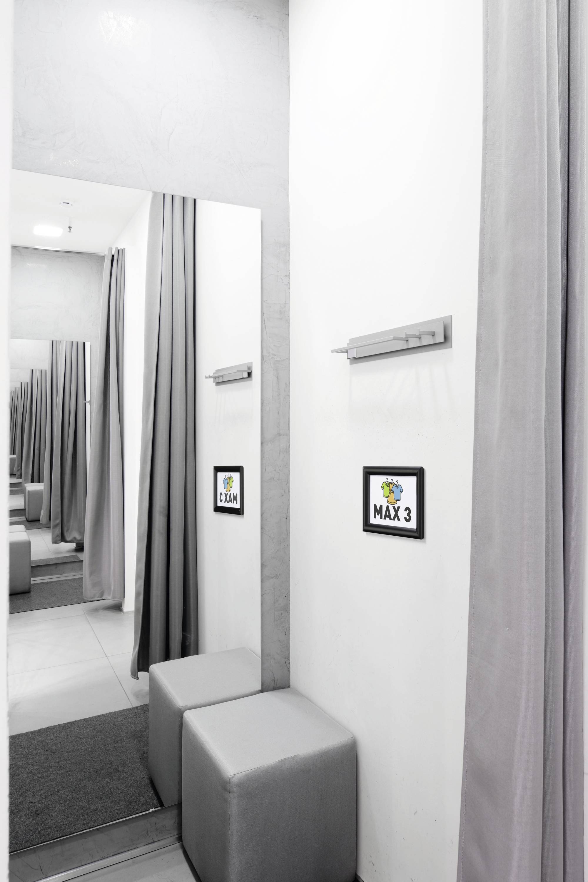 Cabine d'essayage dans un magasin avec un rideau gris et un siège rembourré. Un panneau accroché au mur indique 'MAX 3'.