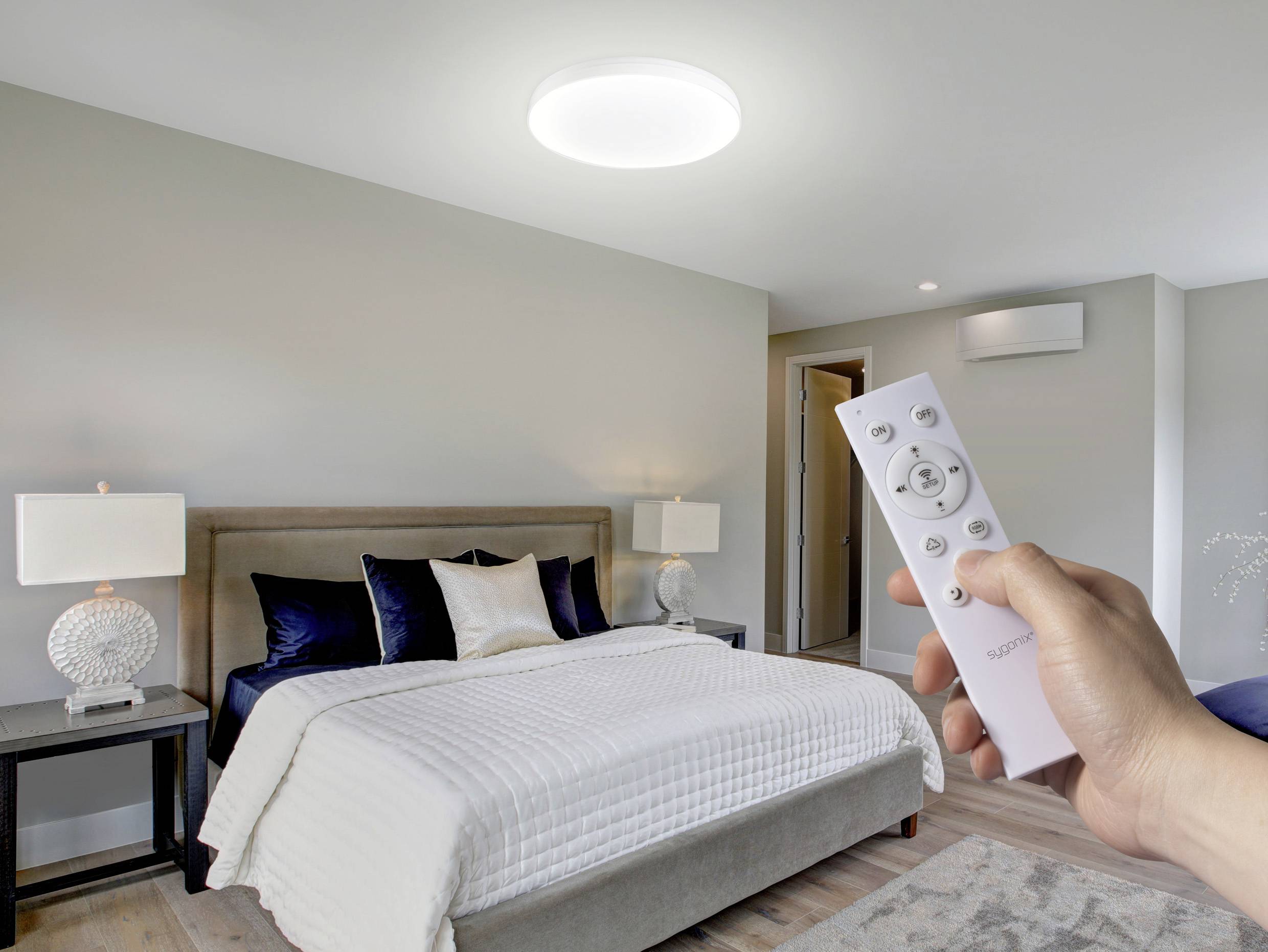 Une chambre moderne avec un lit, des coussins décoratifs et des lampes de chevet. Une main tient une télécommande en direction d'un plafonnier illuminé.