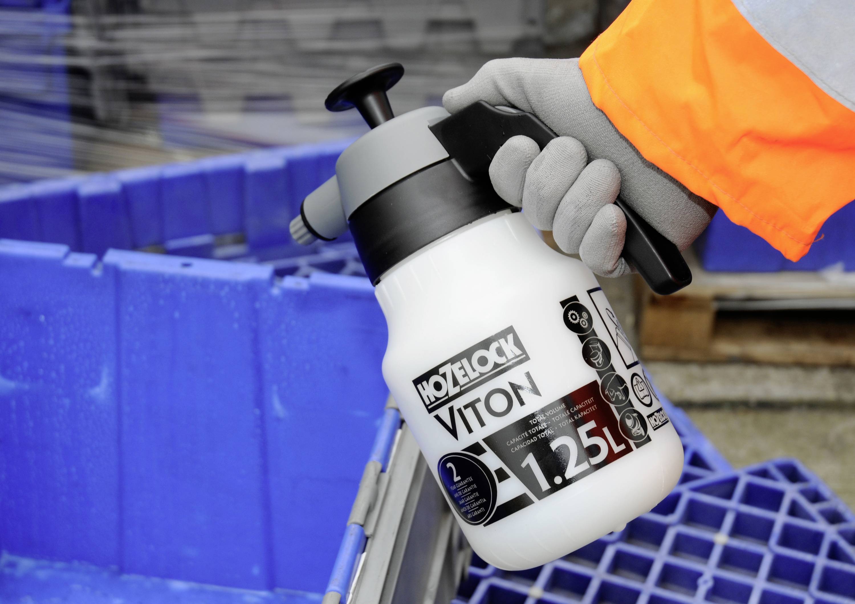 Une main portant des gants de travail tient un récipient de pulvérisation blanc portant l'inscription 'Hozelock Viton 1.25L' devant une palette bleue.