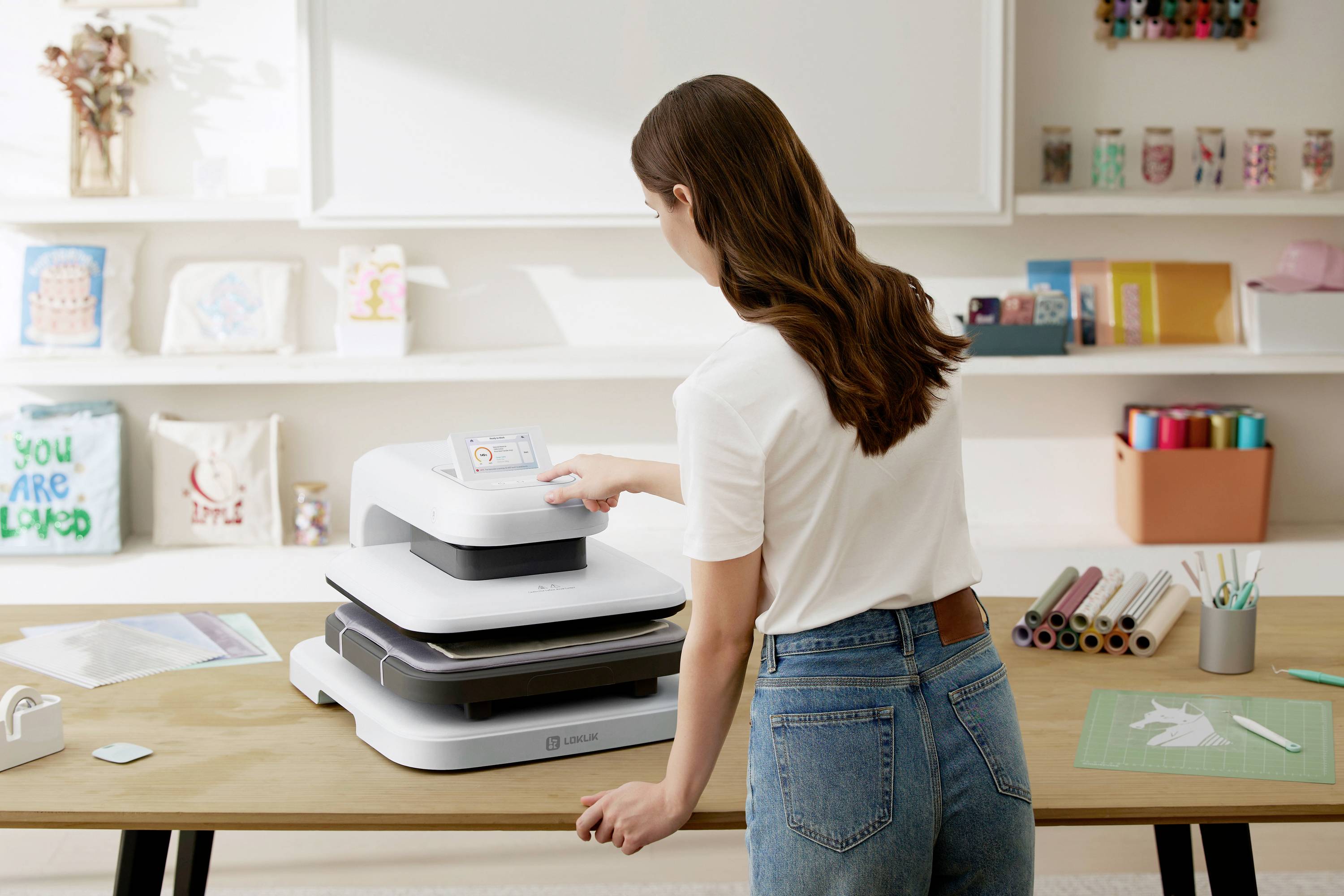 Une femme se tient devant une presse de transfert et en actionne le panneau de commande. Sur la table sont disposés des matériaux de bricolage et des feuilles de transfert.