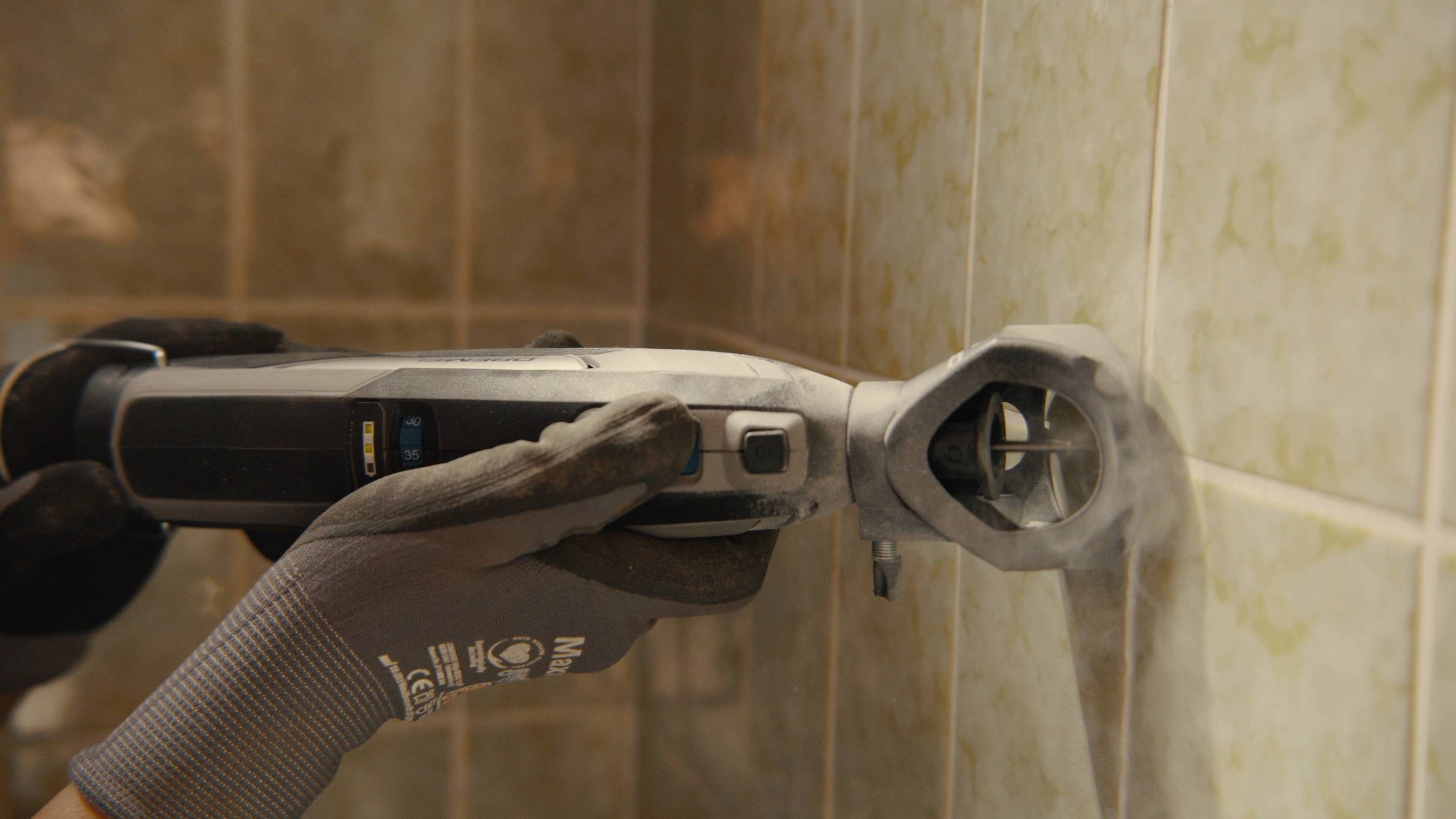 Une personne utilise un outil multifonction électrique pour travailler les joints de carrelage. Des gants de protection sont portés.
