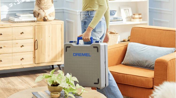 Une personne transportant un coffret d'outils Dremel dans un salon chaleureux et bien éclairé, avec un canapé orange et des meubles en bois, mettant en valeur un espace de travail pour un projet domestique.