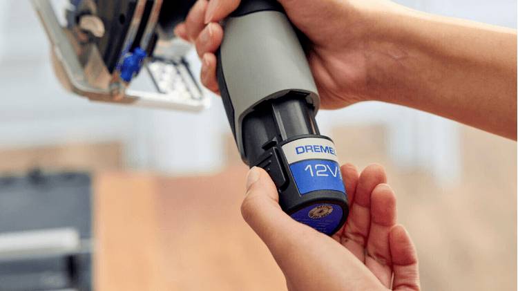 Une personne tient et insère une batterie 'Dremel 12V' bleue et grise dans un outil, en se concentrant sur le processus d'installation de la batterie.