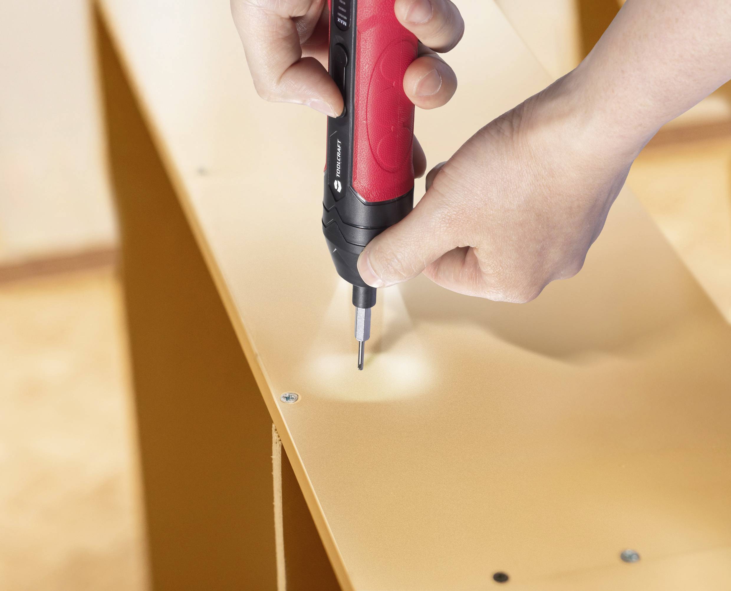 Mains utilisant un tournevis électrique rouge et noir pour assembler un meuble, en vissant des vis dans une surface en bois.