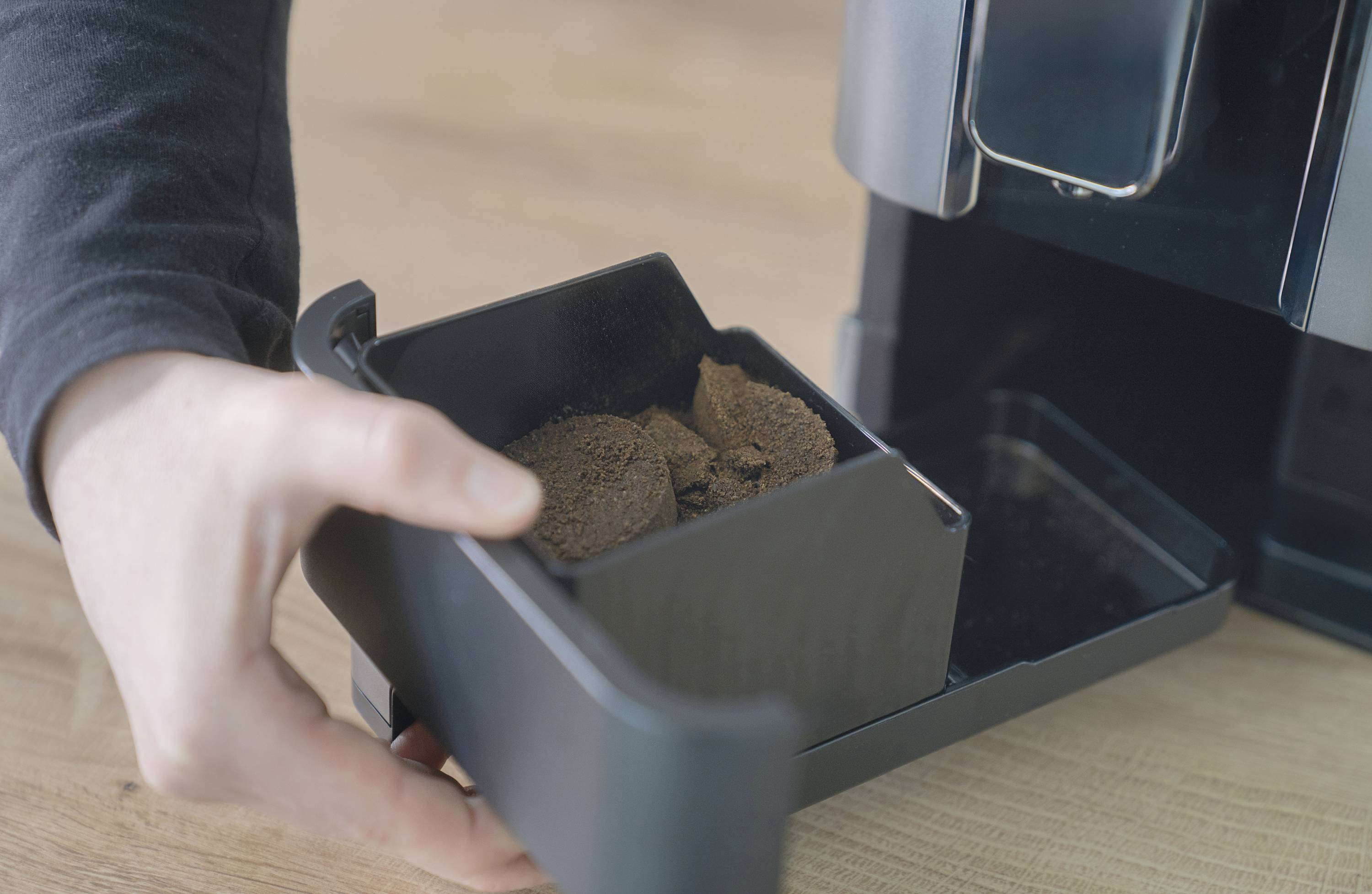 Une main vide le bac à marc de café d'une machine à café sur une surface en bois.