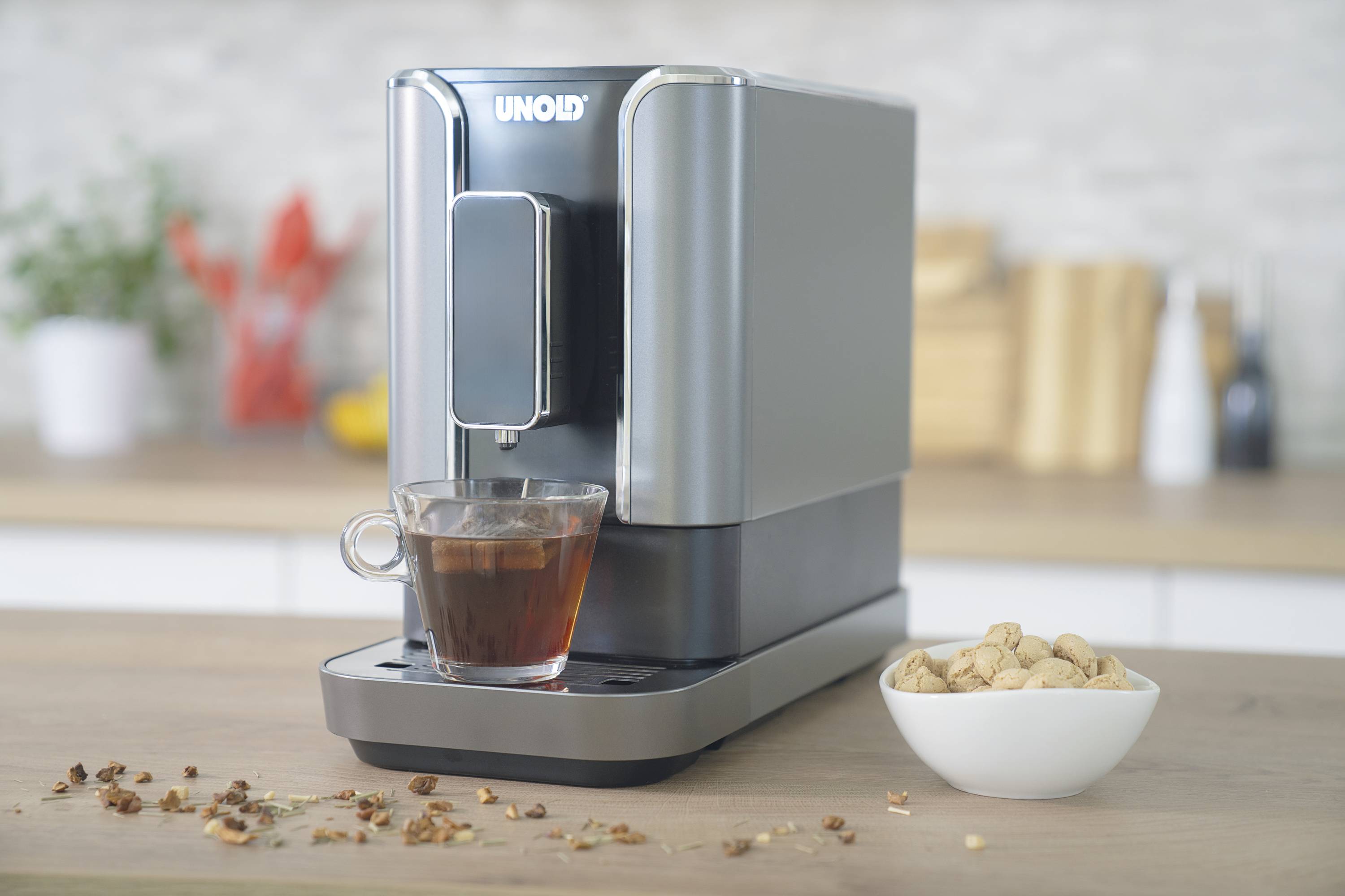 Une machine à café argentée sur un plan de travail de cuisine avec une tasse de thé et un bol de noix à côté. En arrière-plan, des ustensiles de cuisine flous.