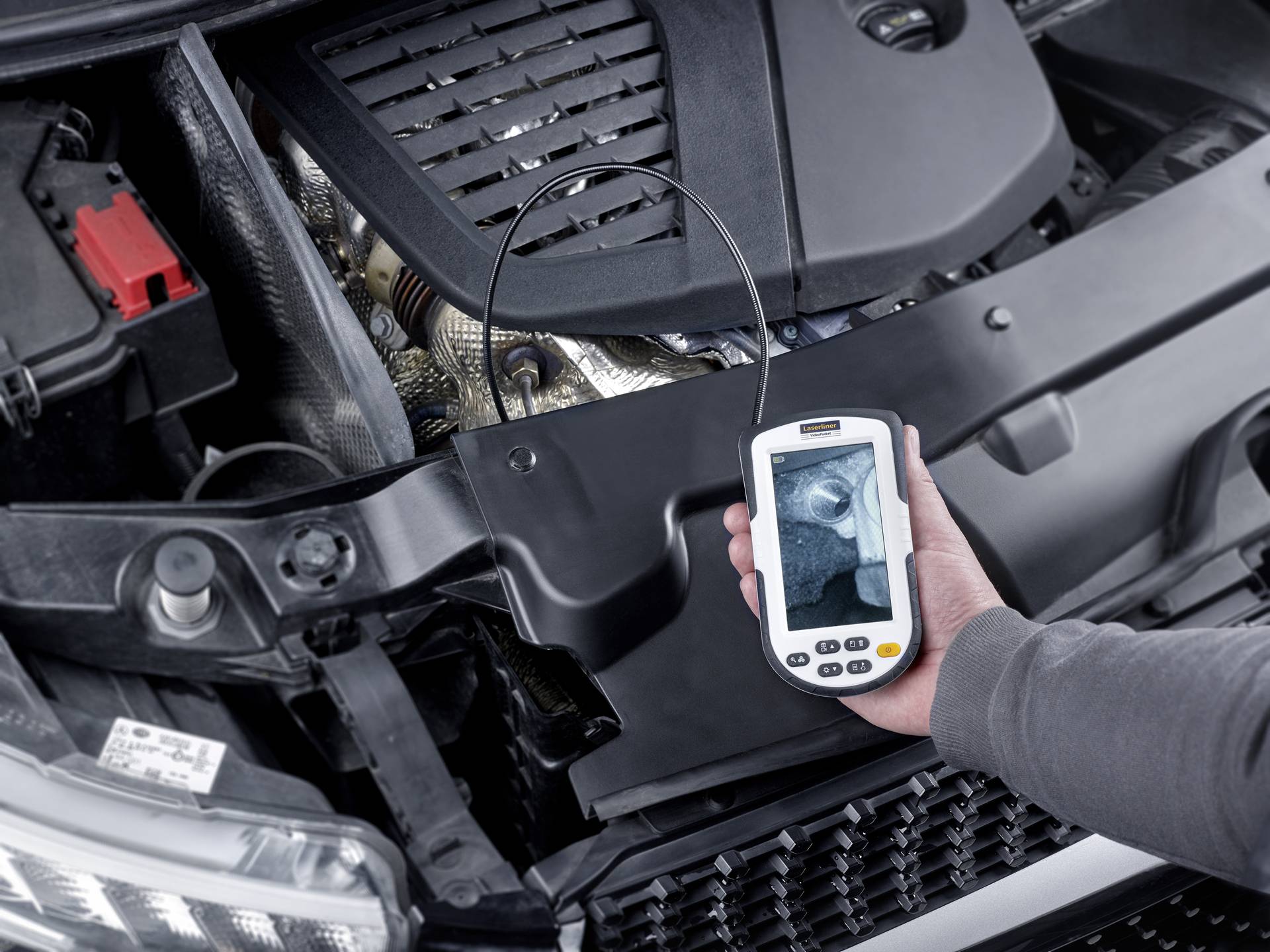 Une personne tient un endoscope qui examine le compartiment moteur d'une voiture. L'écran affiche un gros plan d'une zone du moteur.