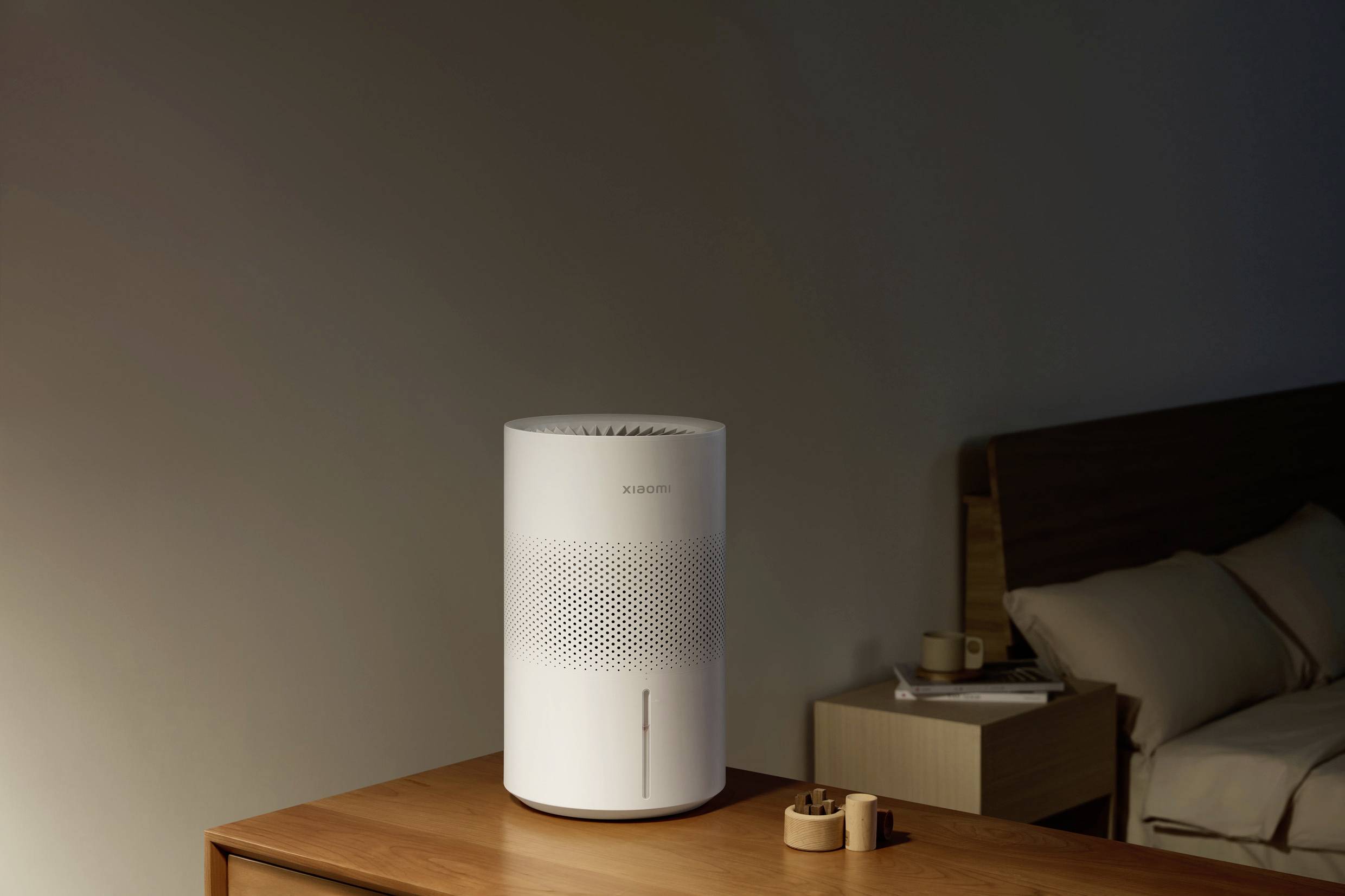Un purificateur d'air blanc est posé sur une table en bois dans une chambre à coucher. Un lit et une table de nuit sont visibles en arrière-plan.