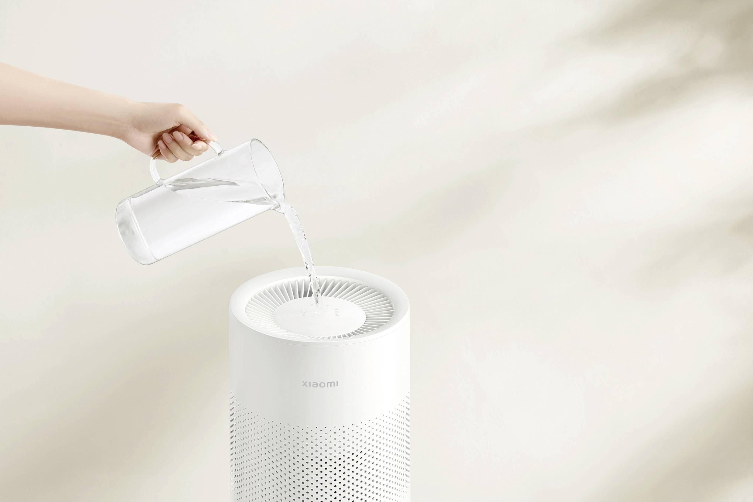 Une main verse de l'eau d'une cruche dans un purificateur d'air. Le purificateur d'air est placé sur un fond neutre.