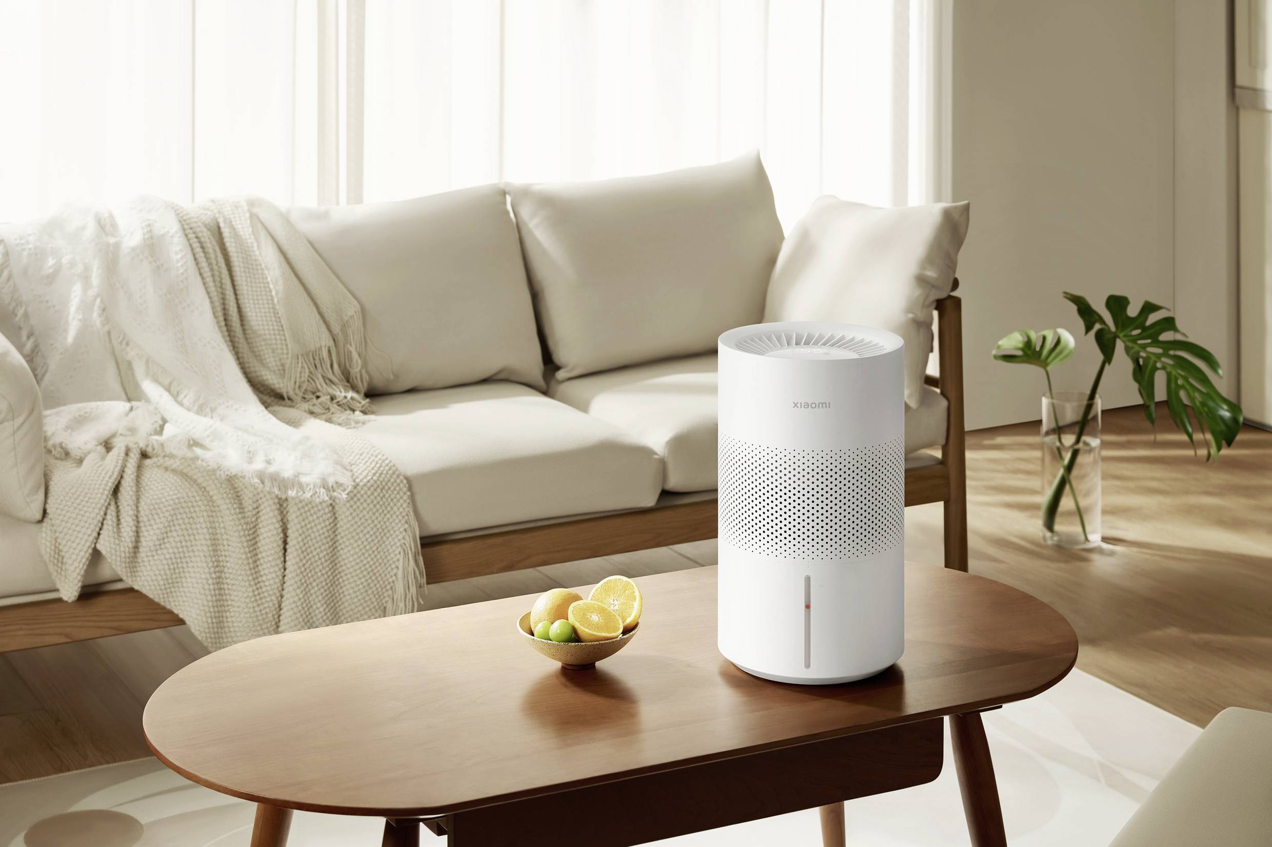 Un purificateur d'air est posé sur une table en bois dans un salon lumineux. En arrière-plan, un canapé avec des coussins et une couverture.