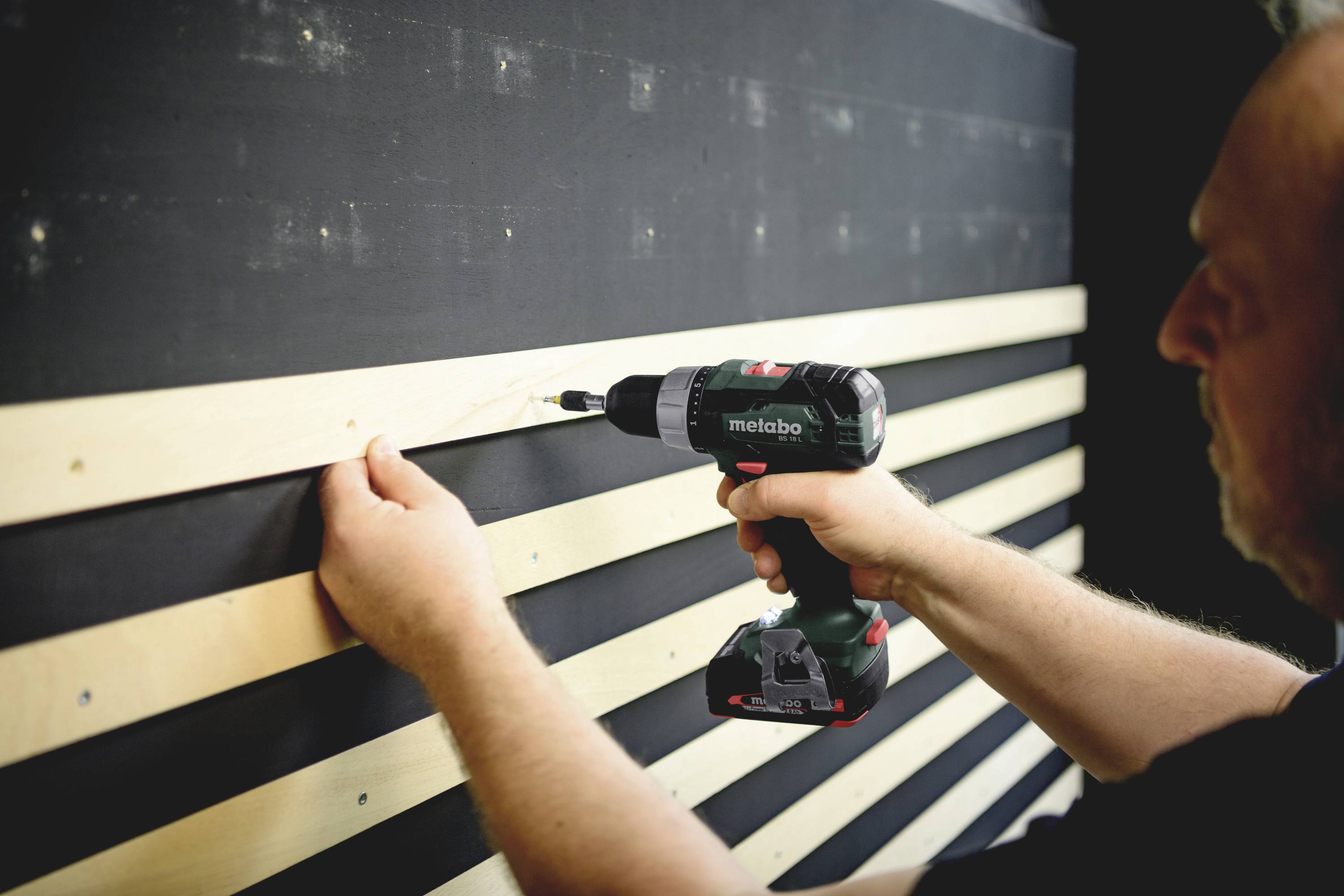 Une personne installe des lattes de bois sur un mur noir à l'aide d'une perceuse sans fil.