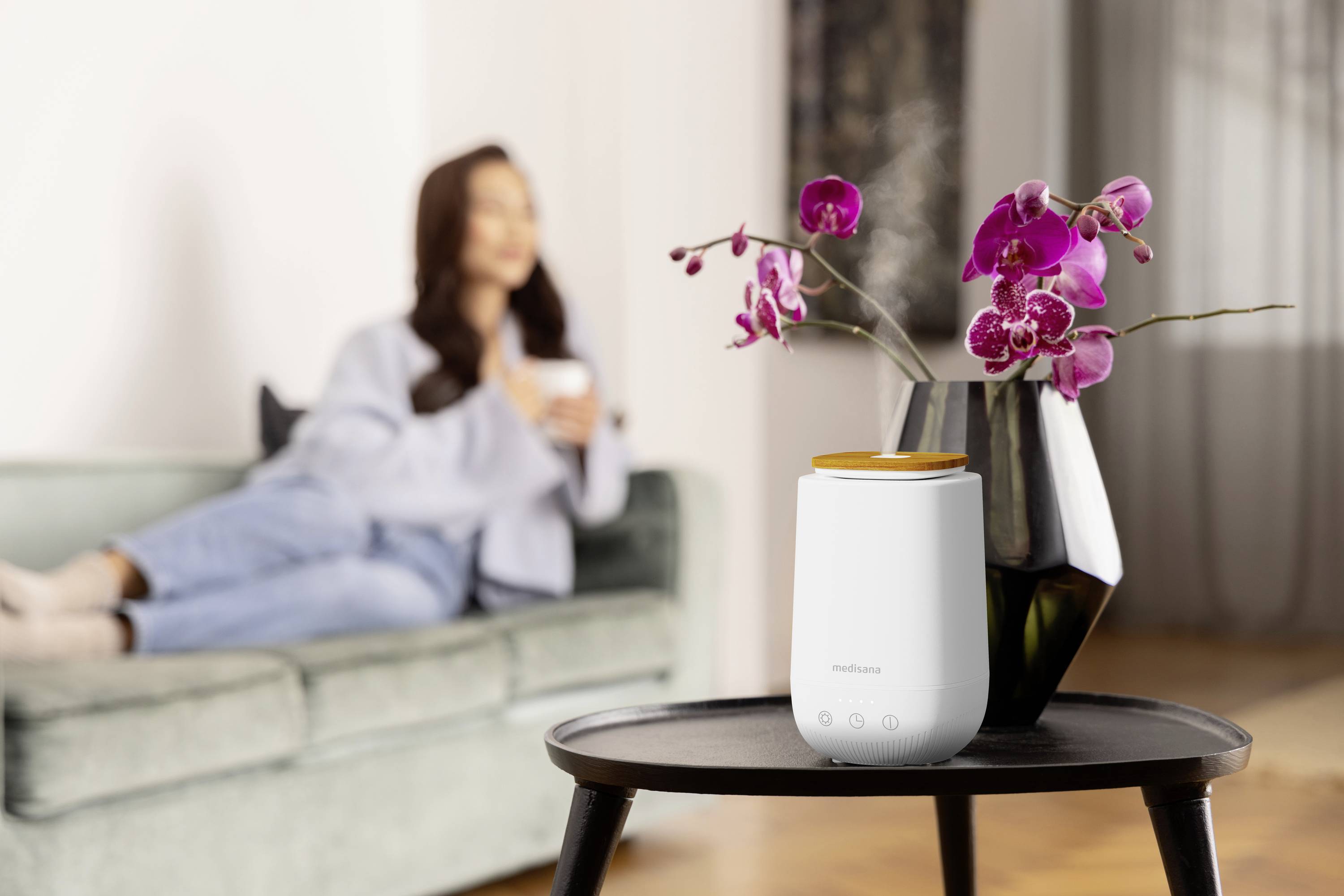 Un purificateur d'air est posé sur une table à côté d'un vase contenant des orchidées mauves. En arrière-plan, une personne floue est assise sur un canapé.