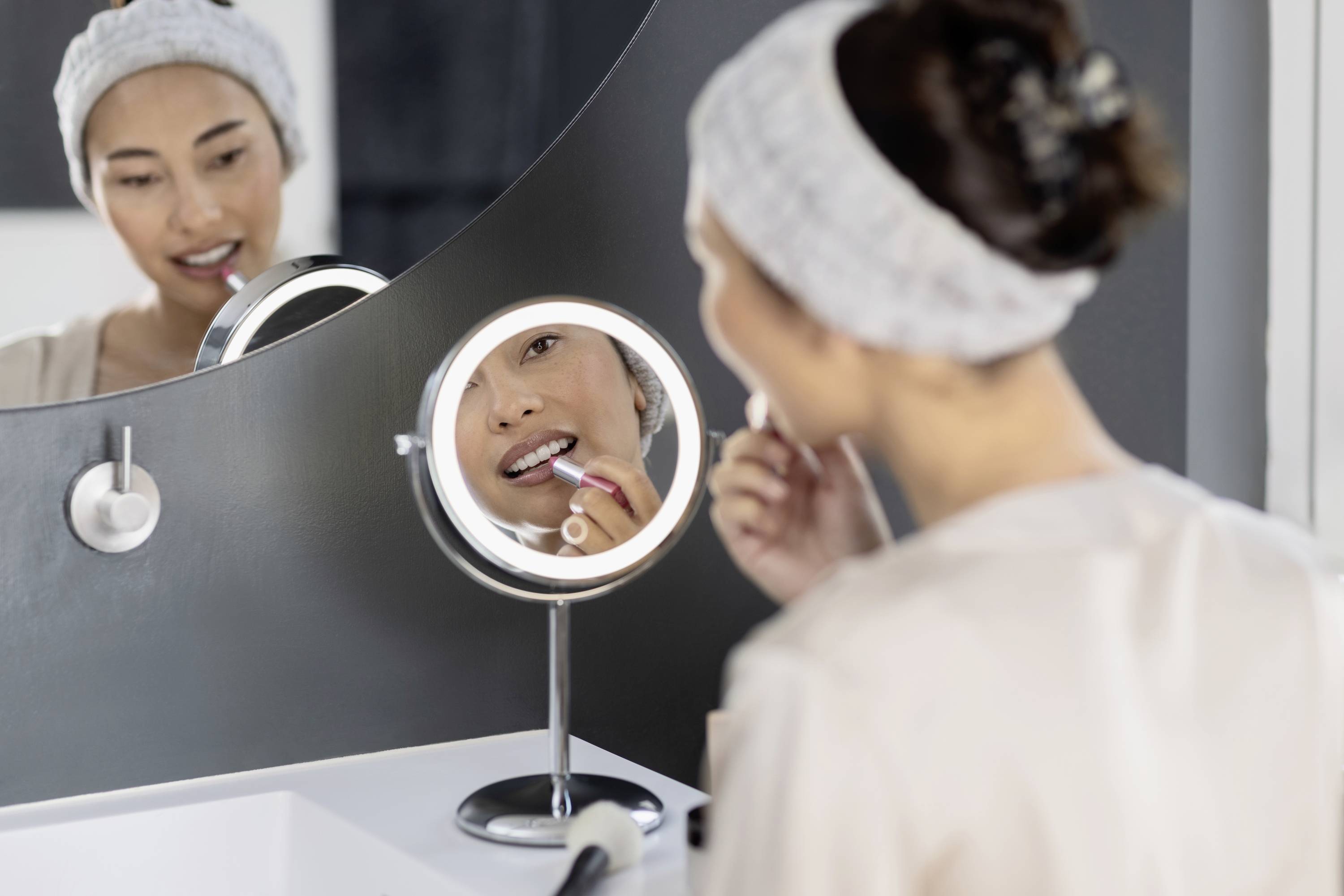 Une femme applique du rouge à lèvres devant un miroir rond dans la salle de bain. Elle porte un bandeau et regarde son reflet dans le miroir.