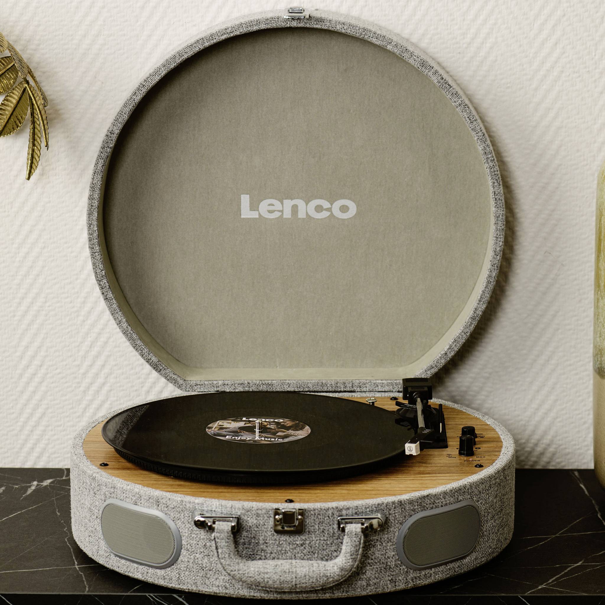 Un tourne-disque gris avec le couvercle ouvert posé sur une table, jouant un disque vinyle. La marque 'Lenco' est visible sur le couvercle.