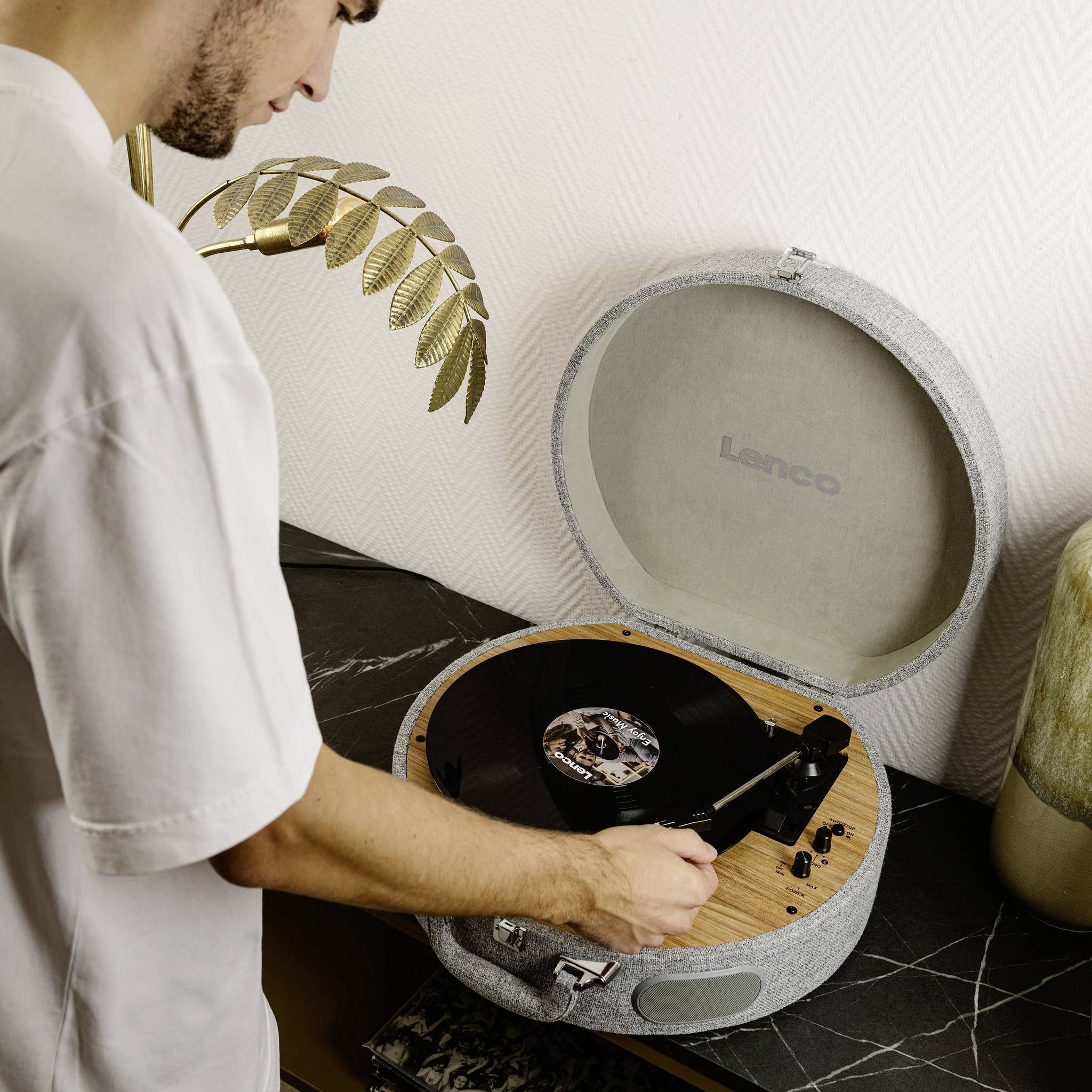 Un homme pose un disque vinyle sur une platine tourne-disque grise. En arrière-plan, une décoration composée de feuilles dorées.