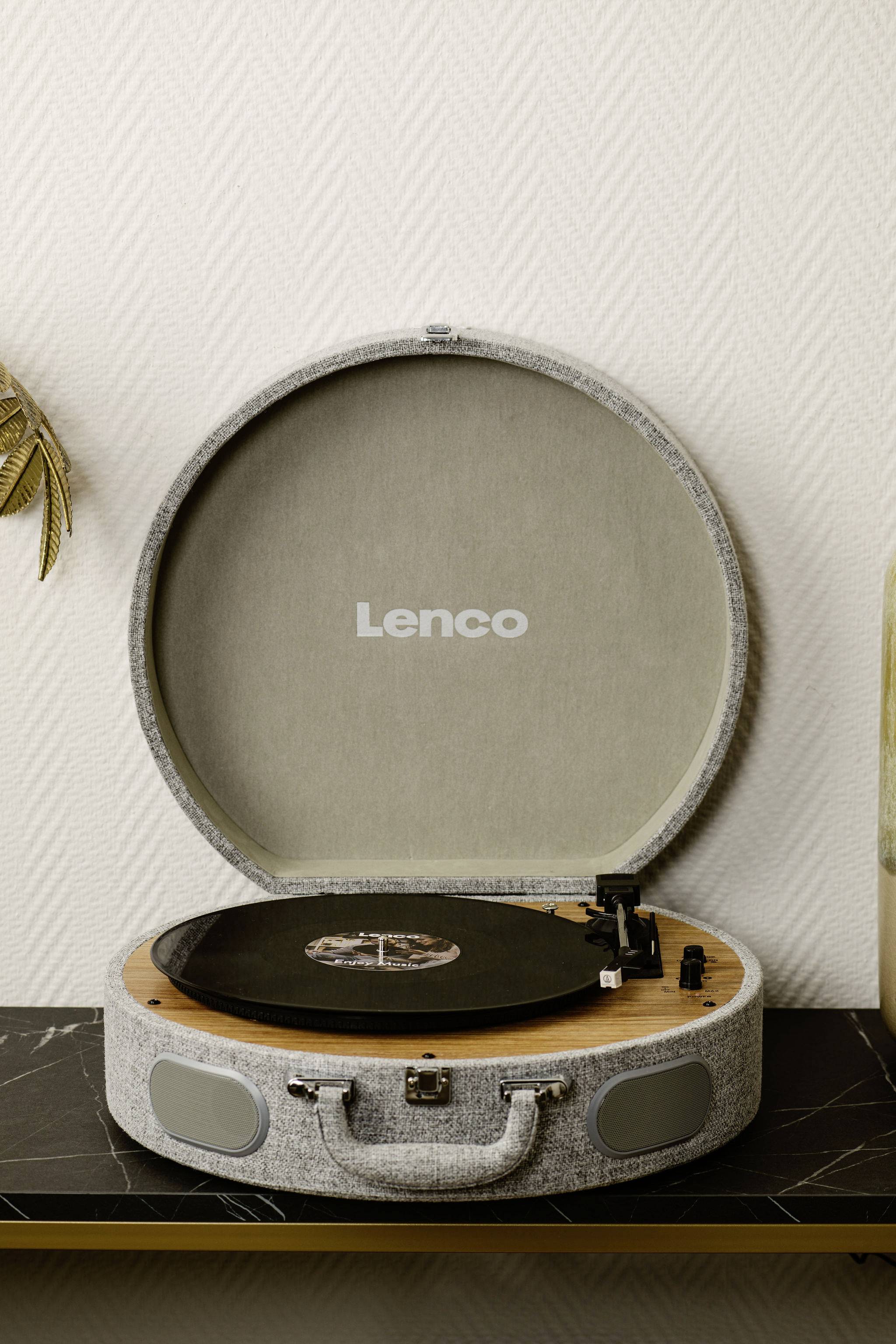 Un tourne-disque ouvert au design de valise, avec un disque vinyle, posé sur une table noire devant un mur texturé.