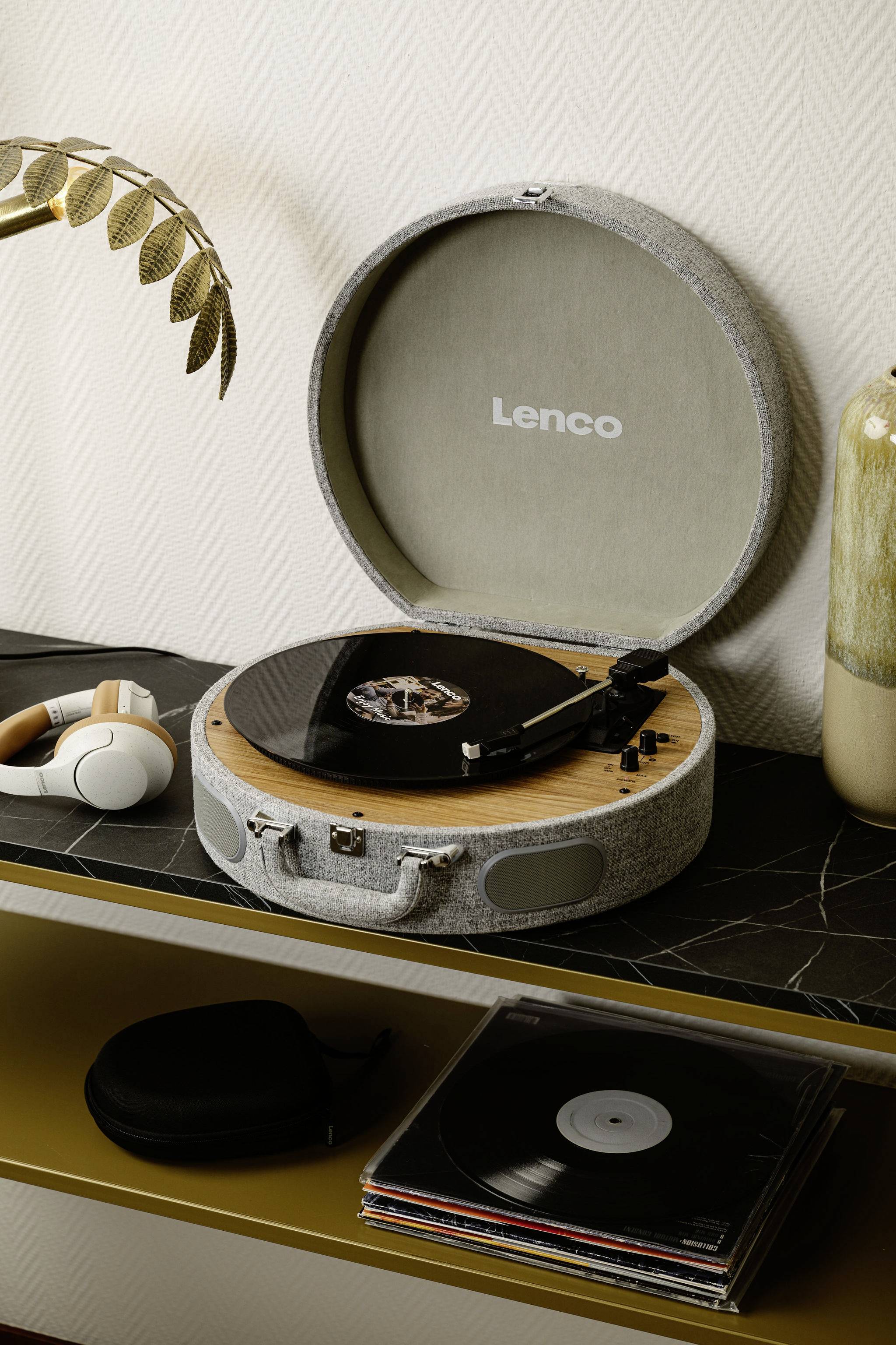 Un tourne-disque ouvert avec un disque vinyle est posé sur une étagère, des écouteurs sont à côté. Une plante se trouve en arrière-plan.