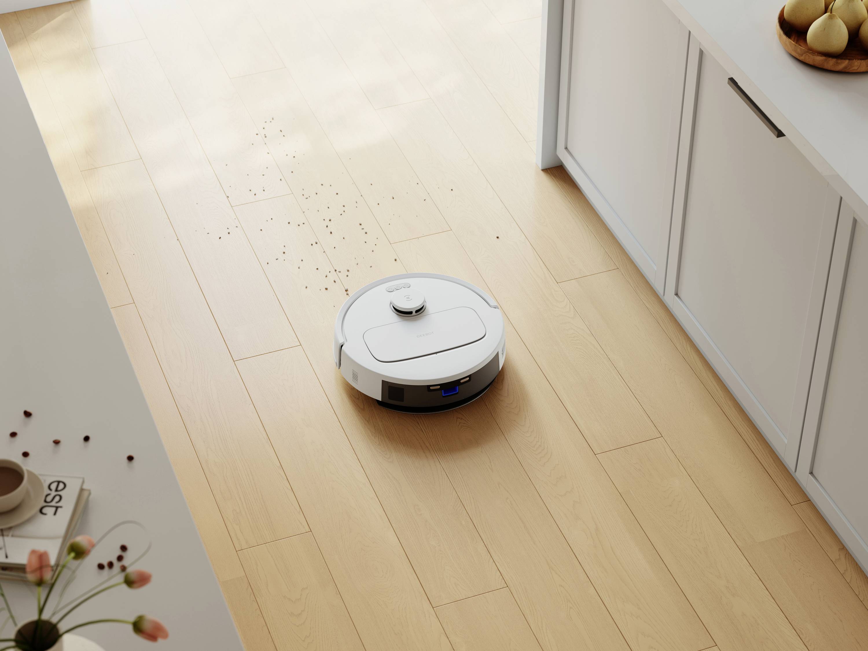 Un robot aspirateur blanc nettoie un sol en bois dans une cuisine où sont éparpillés des miettes. Des placards et un bol de fruits sont visibles en arrière-plan.