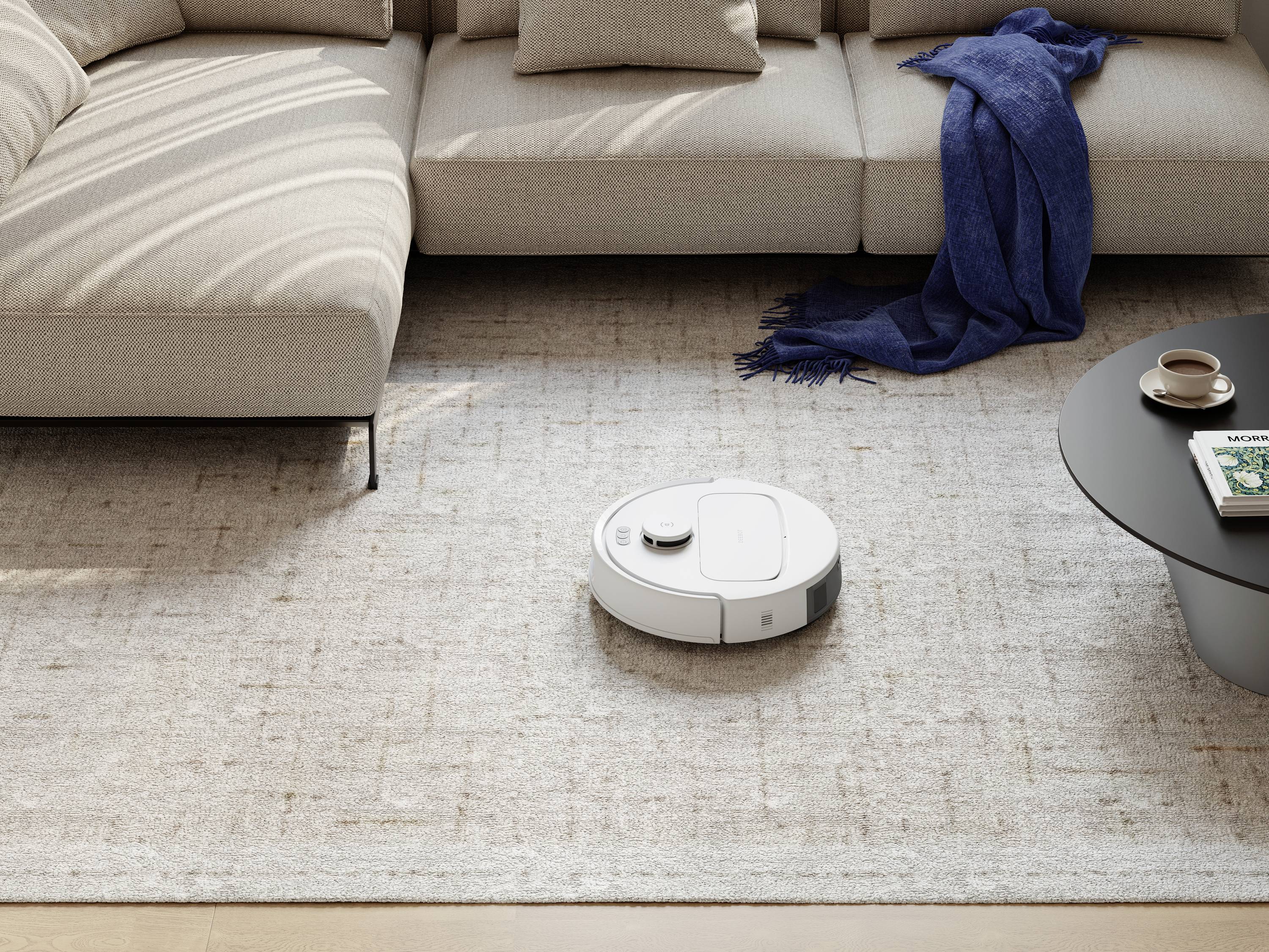 Un robot aspirateur blanc nettoie un tapis beige à côté d'un canapé beige et d'une table basse avec une tasse de café et un foulard bleu.