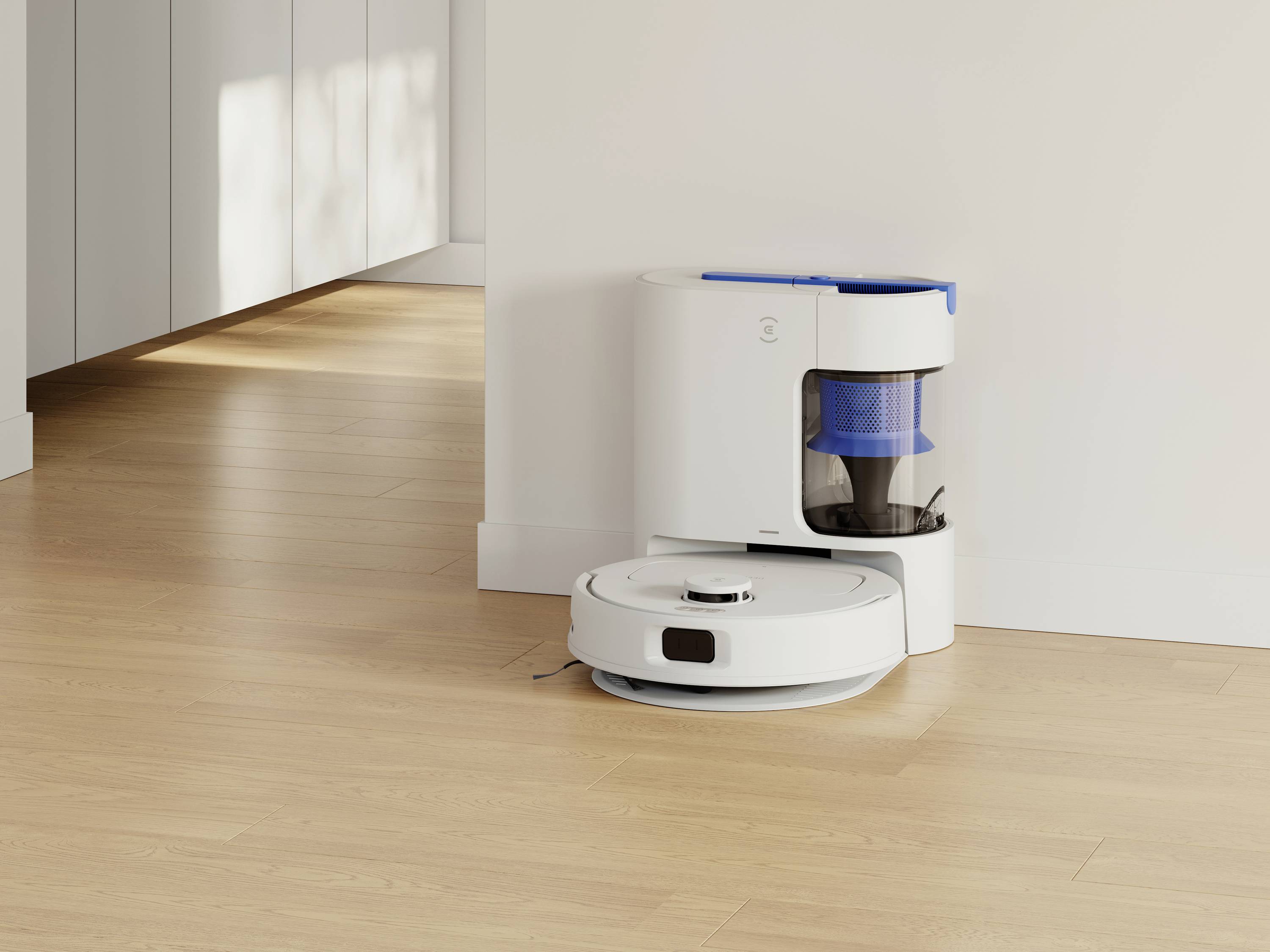 Un robot aspirateur blanc s'arrime à sa station de charge dans une pièce lumineuse au sol en bois. Aucune personne n'est présente dans l'image.
