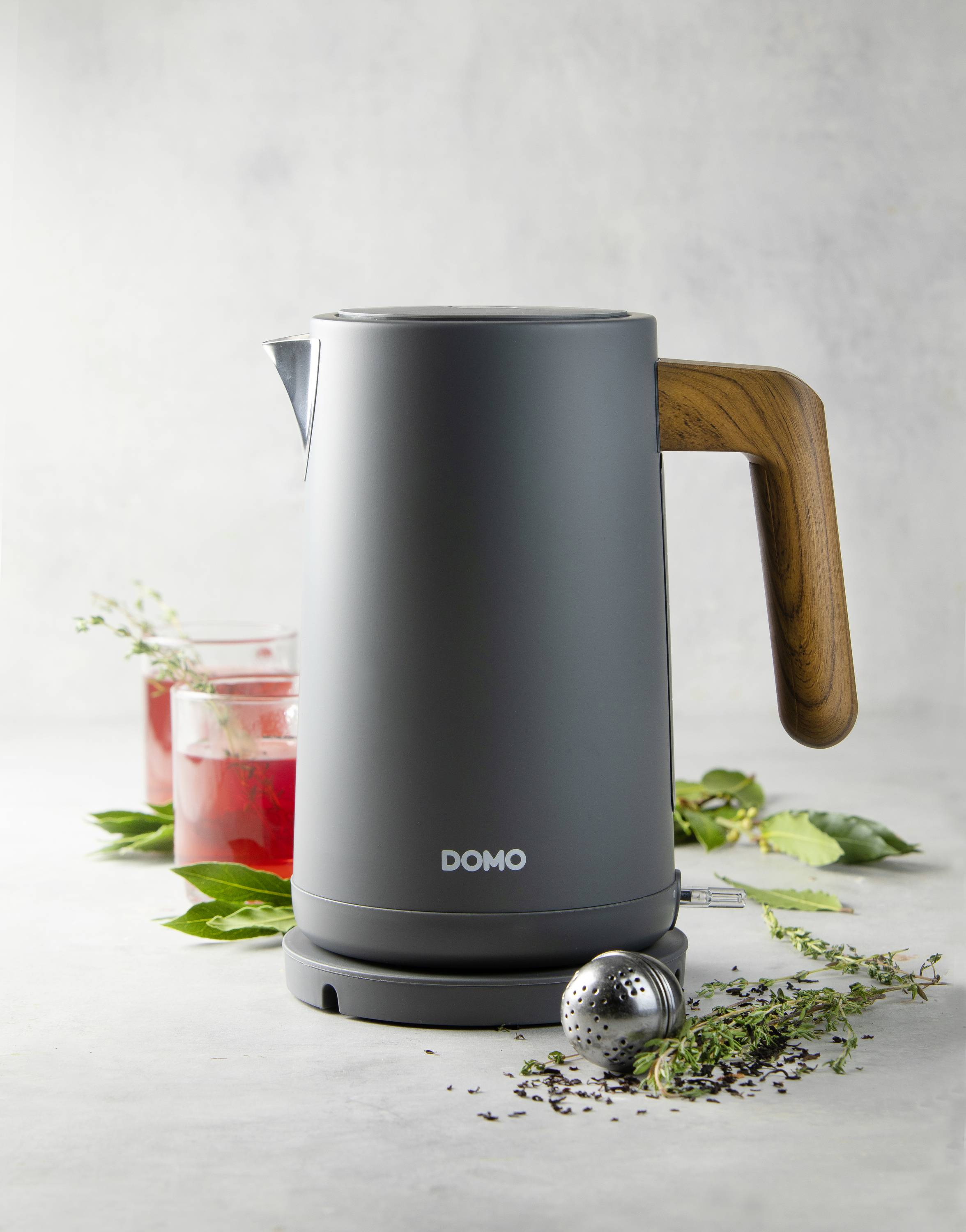 Une bouilloire grise avec une poignée en bois est posée sur une table à côté d'un verre contenant une boisson rouge et des herbes. Marquage : 'DOMO'.
